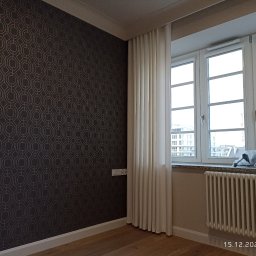 Andrzej Baldin metr.wow.pl - Pokój po odświeżeniu: ciemna ściana z geometrycznym wzorem, białe zasłony przy oknie z widokiem na miasto, grzejnik pod oknem, drewniana podłoga.