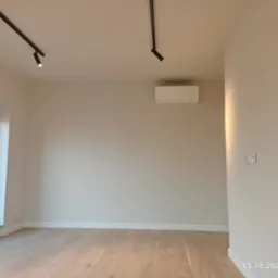 Pusty pokój po remoncie z drewnianą podłogą, świeżo pomalowanymi ścianami w odcieniach beżu, oświetleniem punktowym na szynoprzewodach i klimatyzacją na ścianie.