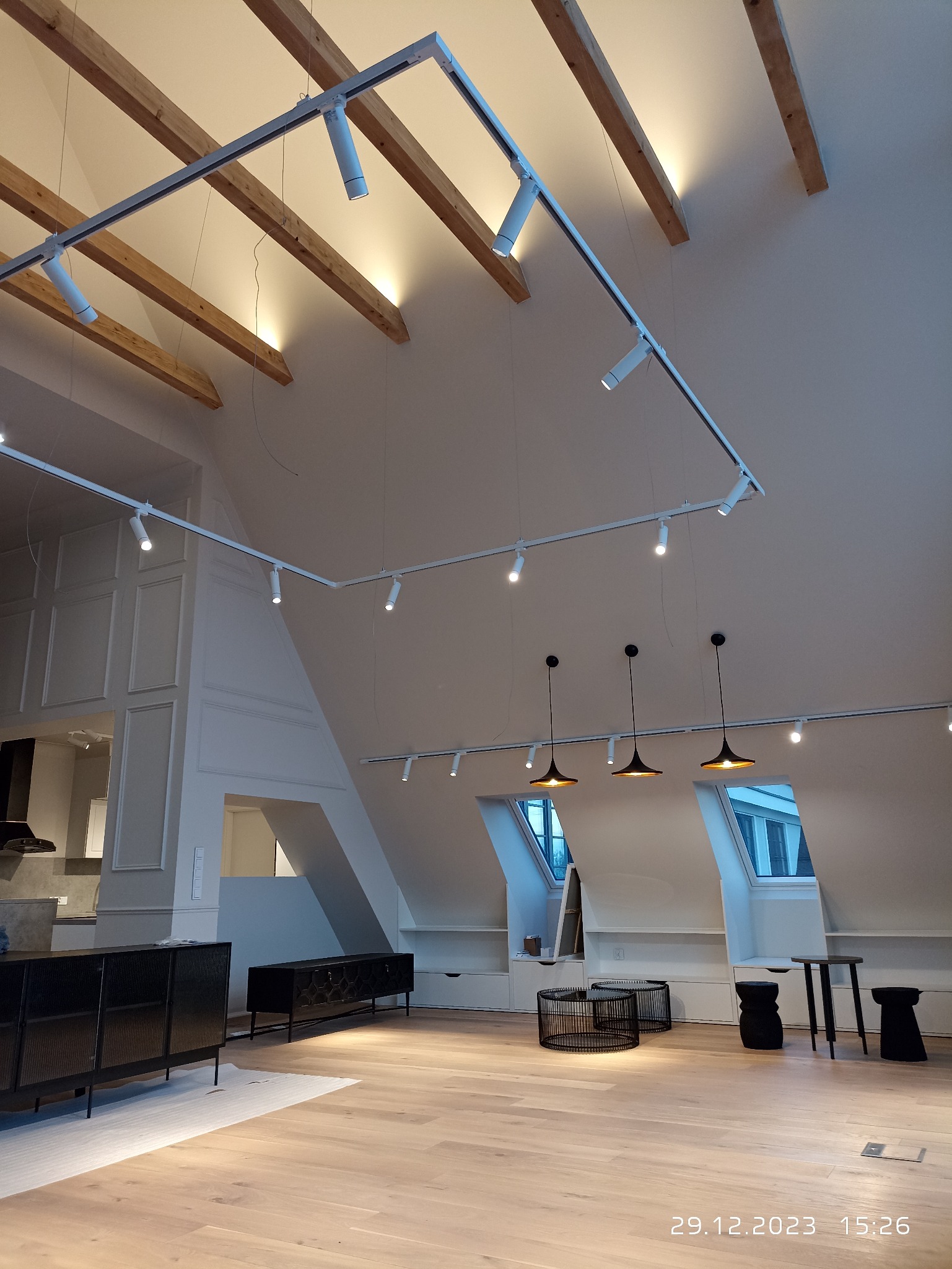 Nowoczesne poddasze po remoncie z drewnianą podłogą, białymi ścianami, drewnianymi belkami stropowymi, oświetleniem szynowym i wiszącymi lampami. W tle widoczna kuchnia.