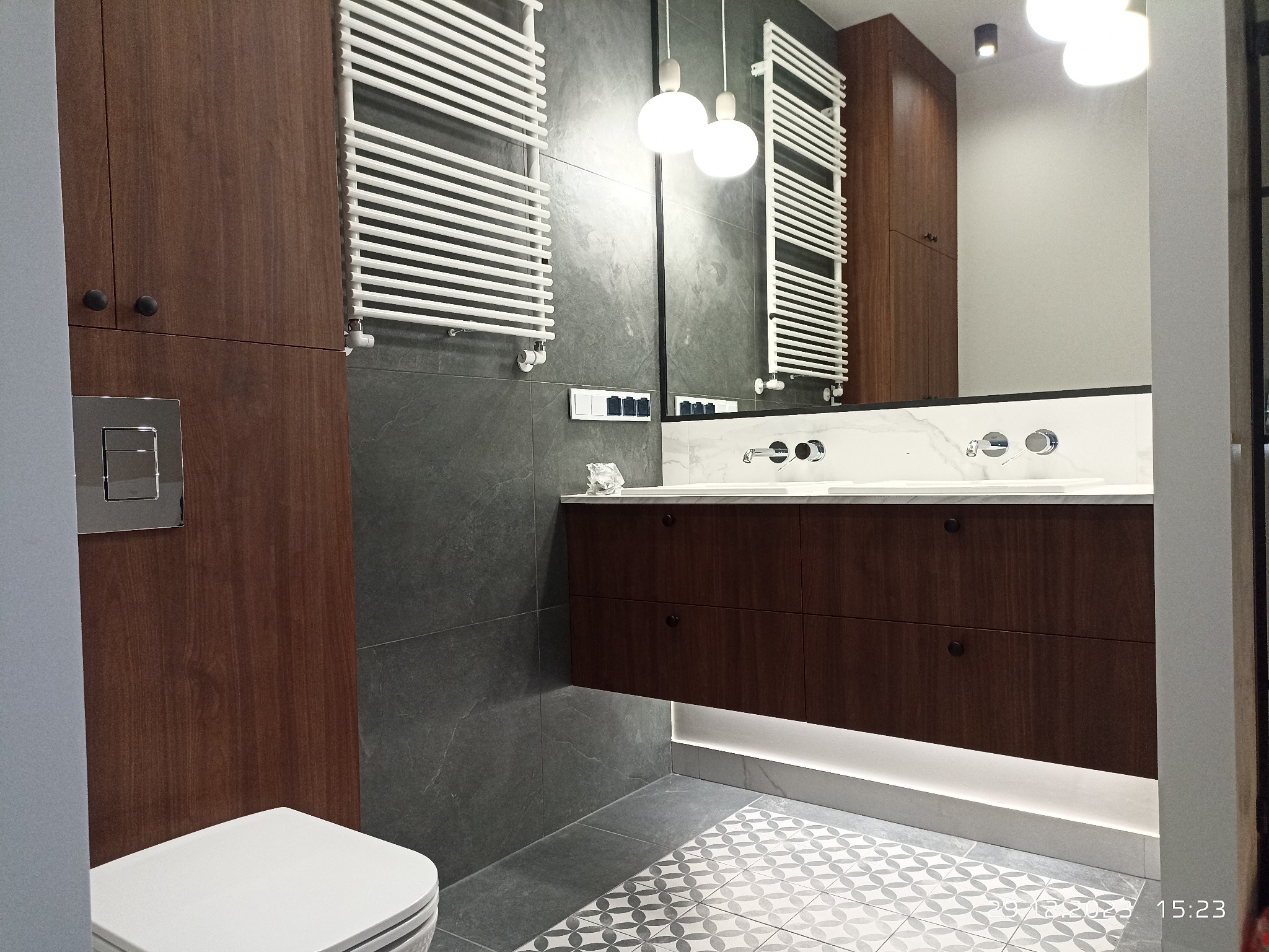 Nowoczesna łazienka z szarą płytką imitującą kamień, wiszącą szafką z umywalkami nablatowymi, grzejnikiem drabinkowym i podświetleniem LED pod szafką.