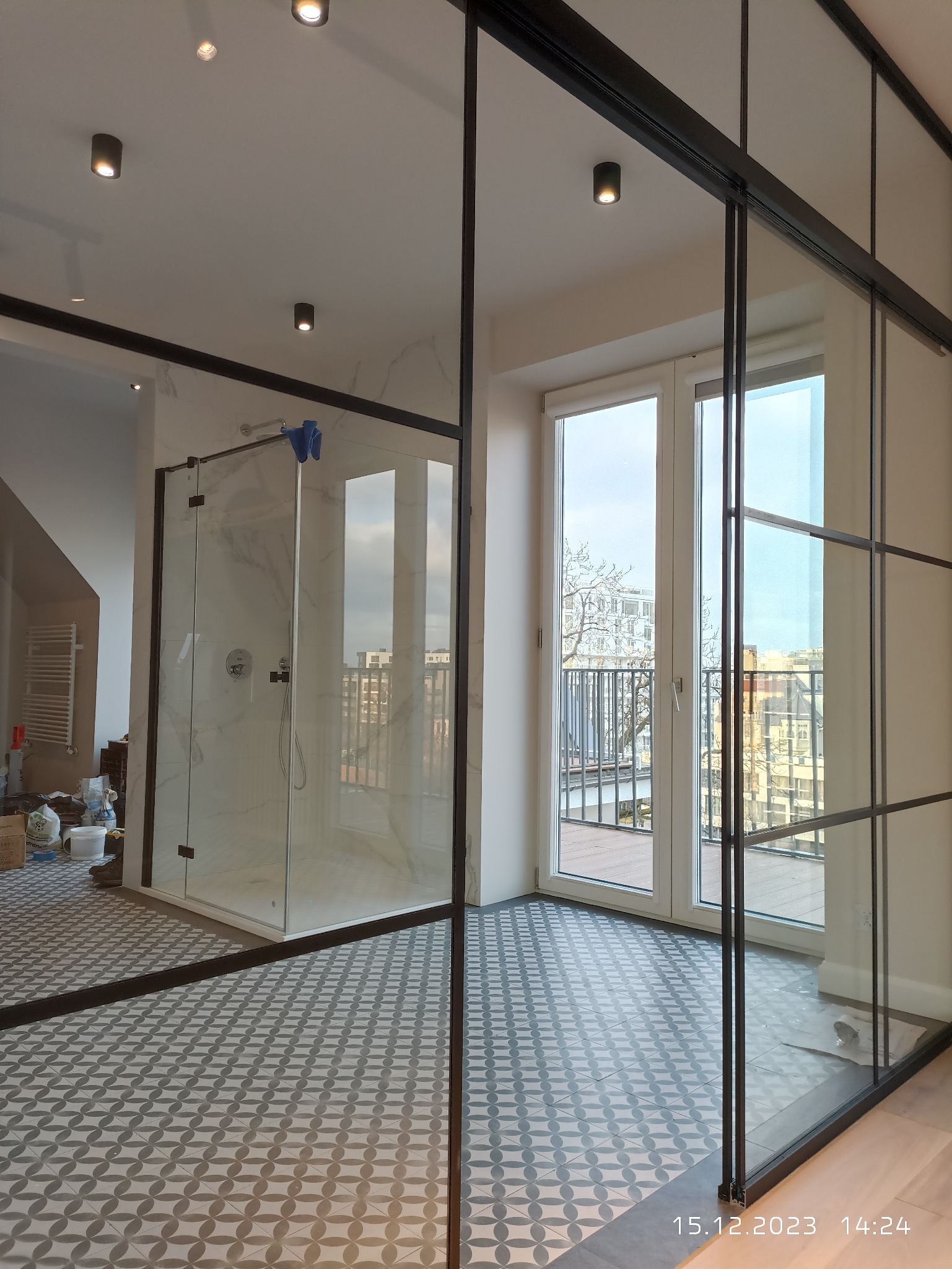 Nowoczesna łazienka z kabiną prysznicową typu walk-in, wykończona marmurową płytką, oddzielona od reszty pomieszczenia szklaną ścianką z czarnymi profilami, widoczne pozostałości po pracach...