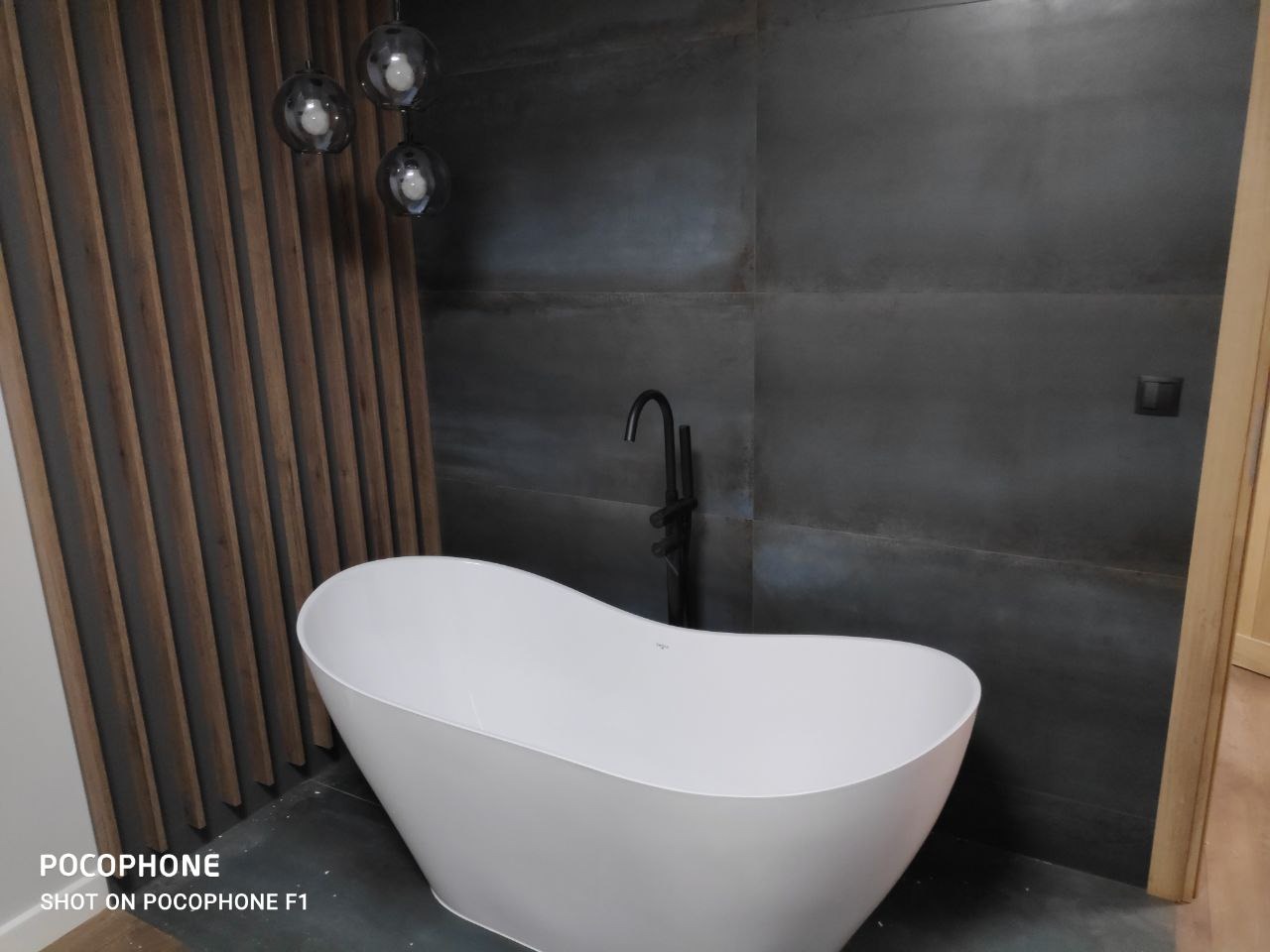 Nowoczesna łazienka z wolnostojącą białą wanną na tle ciemnych, metalicznych płytek i drewnianych lameli, oświetlona wiszącymi lampami kulami.