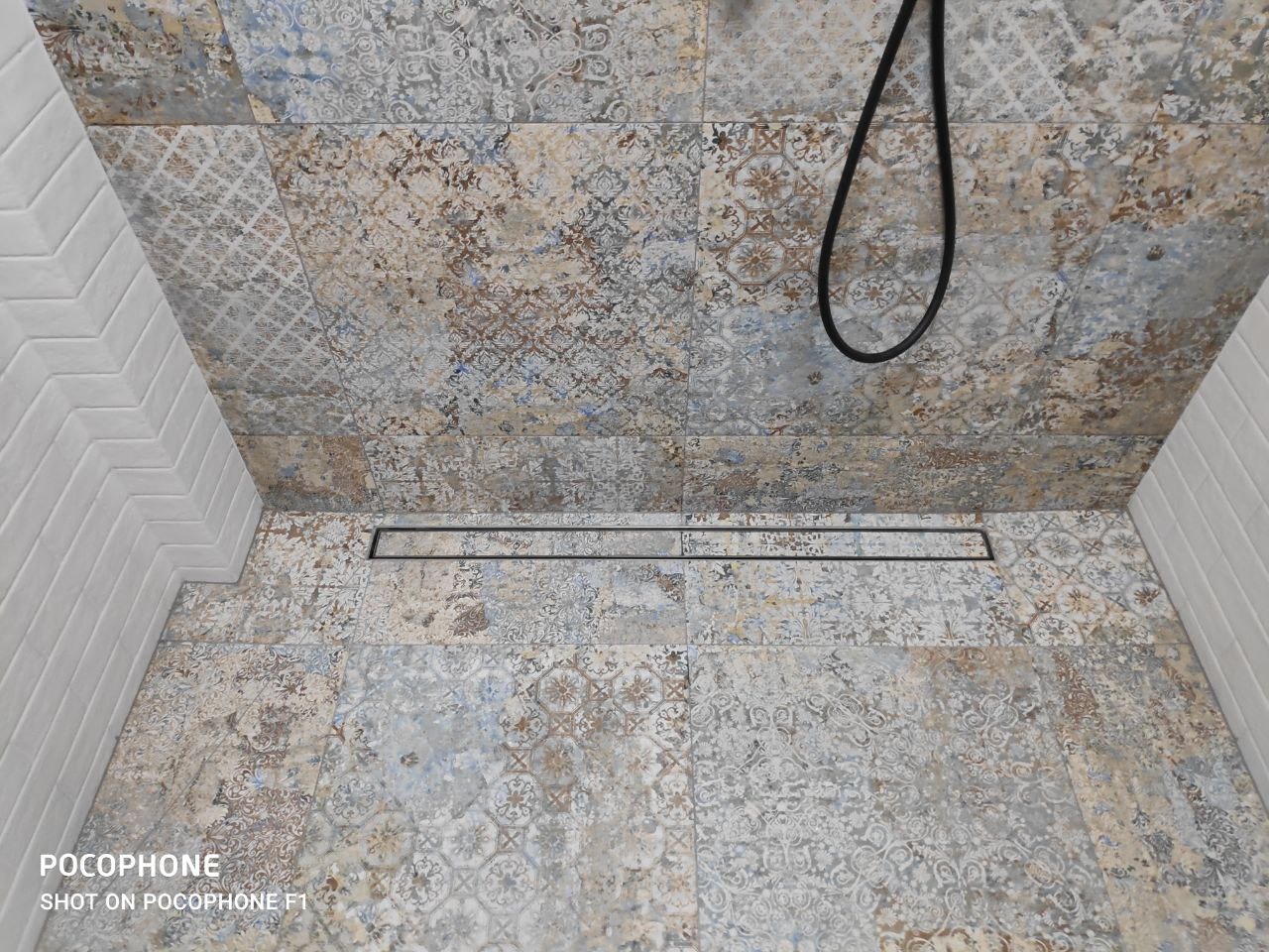 Ukończony odpływ liniowy w kabinie prysznicowej wyłożonej patchworkowymi płytkami w odcieniach beżu i błękitu, białe płytki imitujące cegłę na ścianie, widoczny fragment słuchawki prysznicowej.