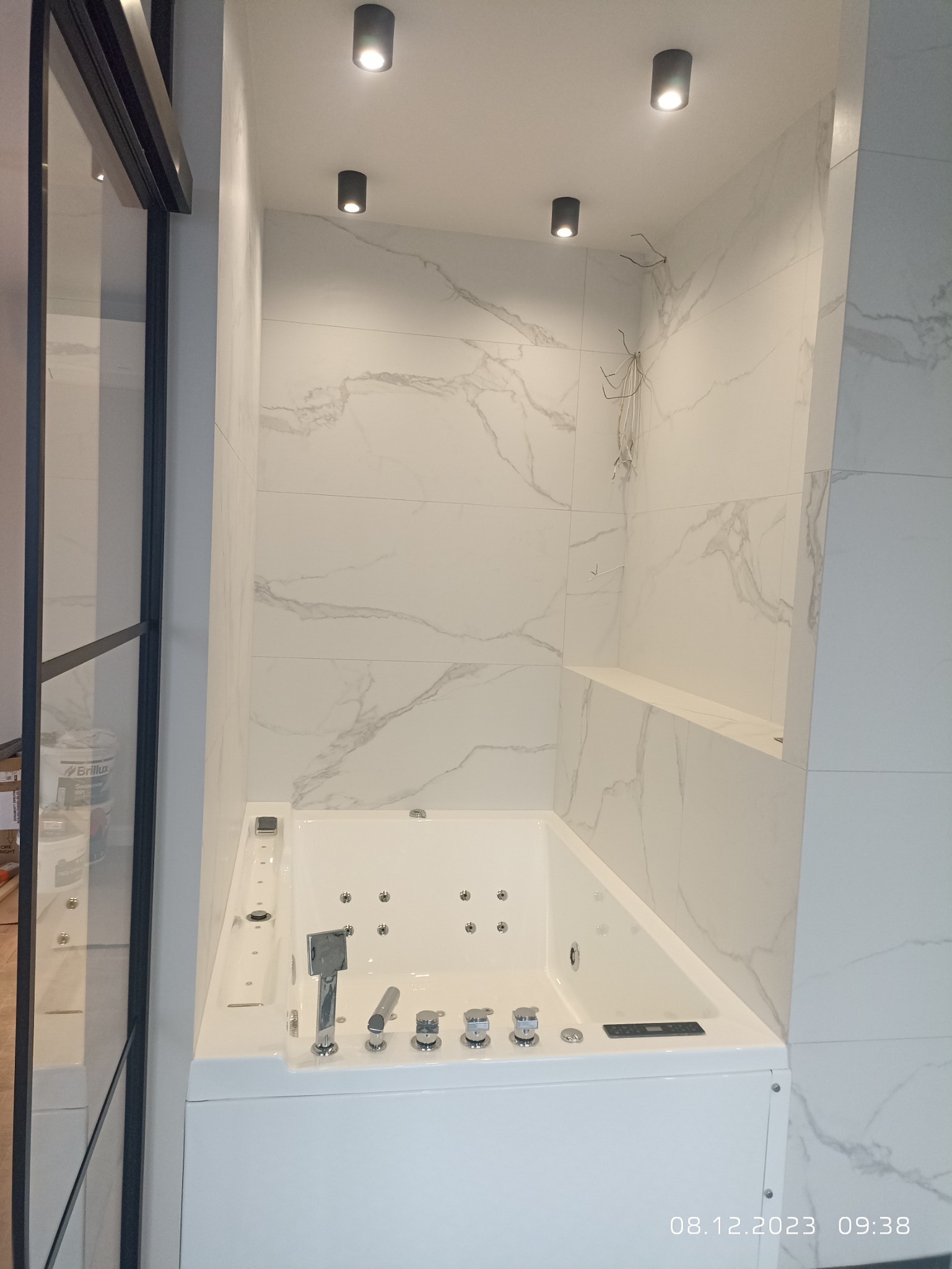 Nowoczesna, narożna wanna z hydromasażem, obudowana białą zabudową, z armaturą w chromowanym wykończeniu, na tle ściany wyłożonej białymi płytami imitującymi marmur, z widocznymi przewodami...