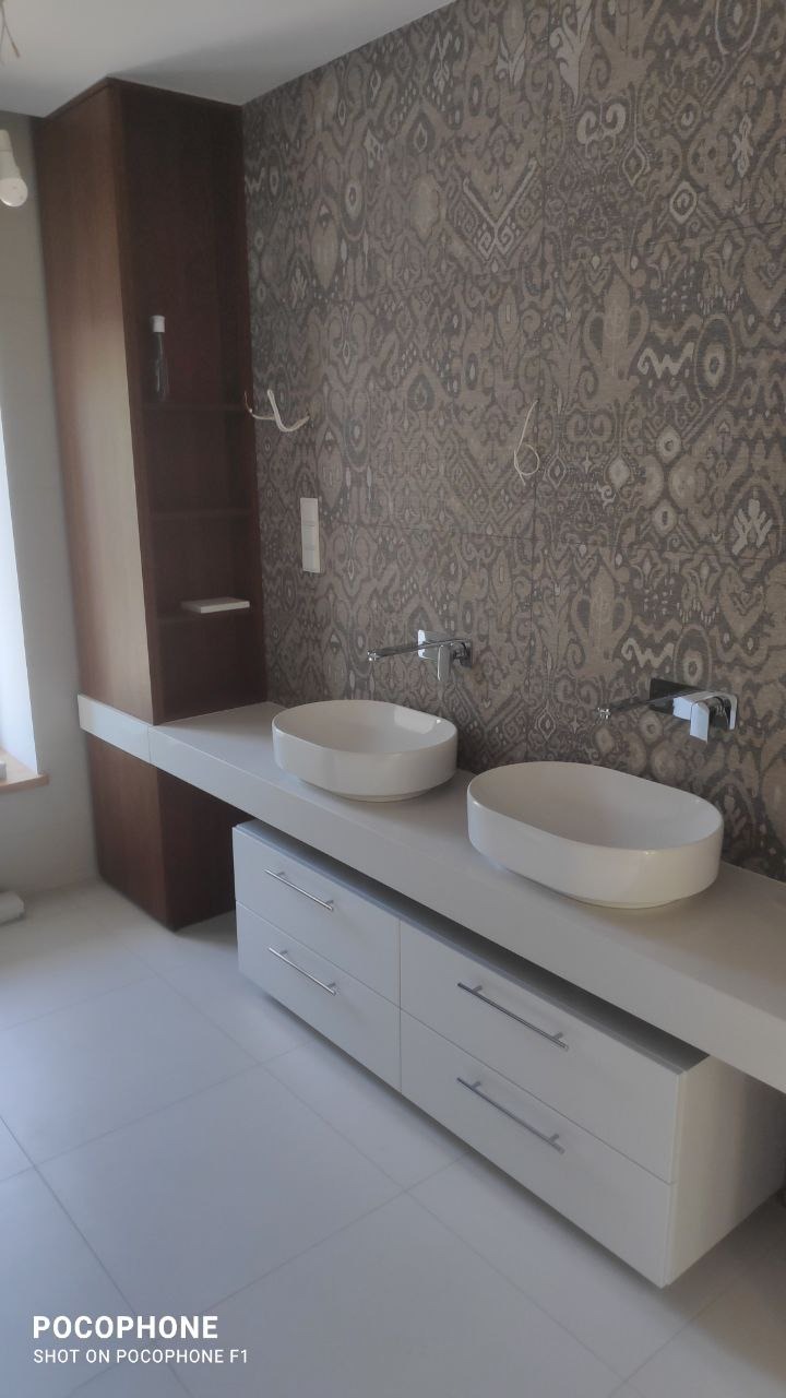 Nowoczesna łazienka z dwiema owalnymi umywalkami nablatowymi, białymi szafkami z poziomymi uchwytami, brązową szafką z półkami i ścianą pokrytą wzorzystą tapetą w odcieniach szarości i beżu.