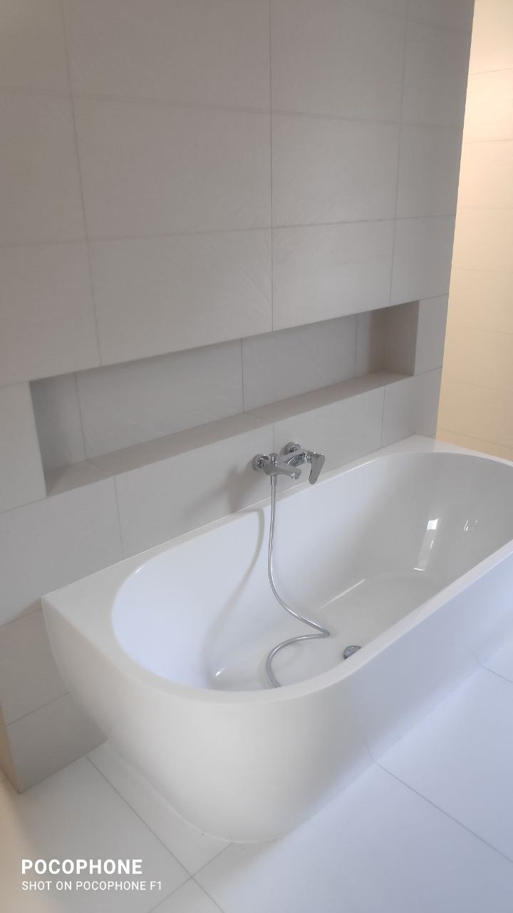 Minimalistyczna, biała łazienka z wanną, chromowaną baterią i wnęką w ścianie wyłożonej płytkami w odcieniach bieli.