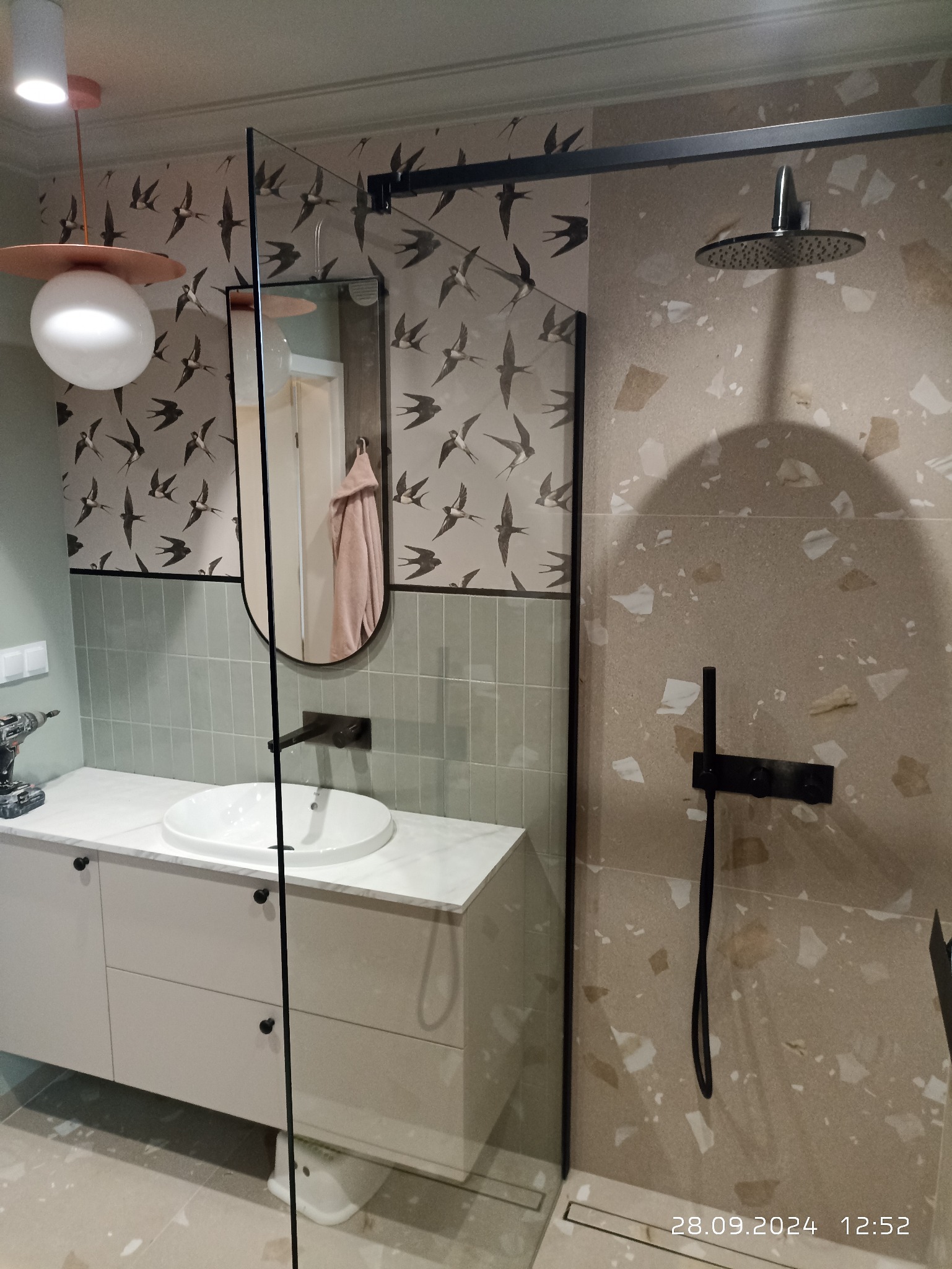 Nowoczesna łazienka z kabiną prysznicową typu walk-in, umywalką nablatową na szafce z szufladami, lustrem w czarnej ramie i tapetą w jaskółki. Na blacie wiertarka.