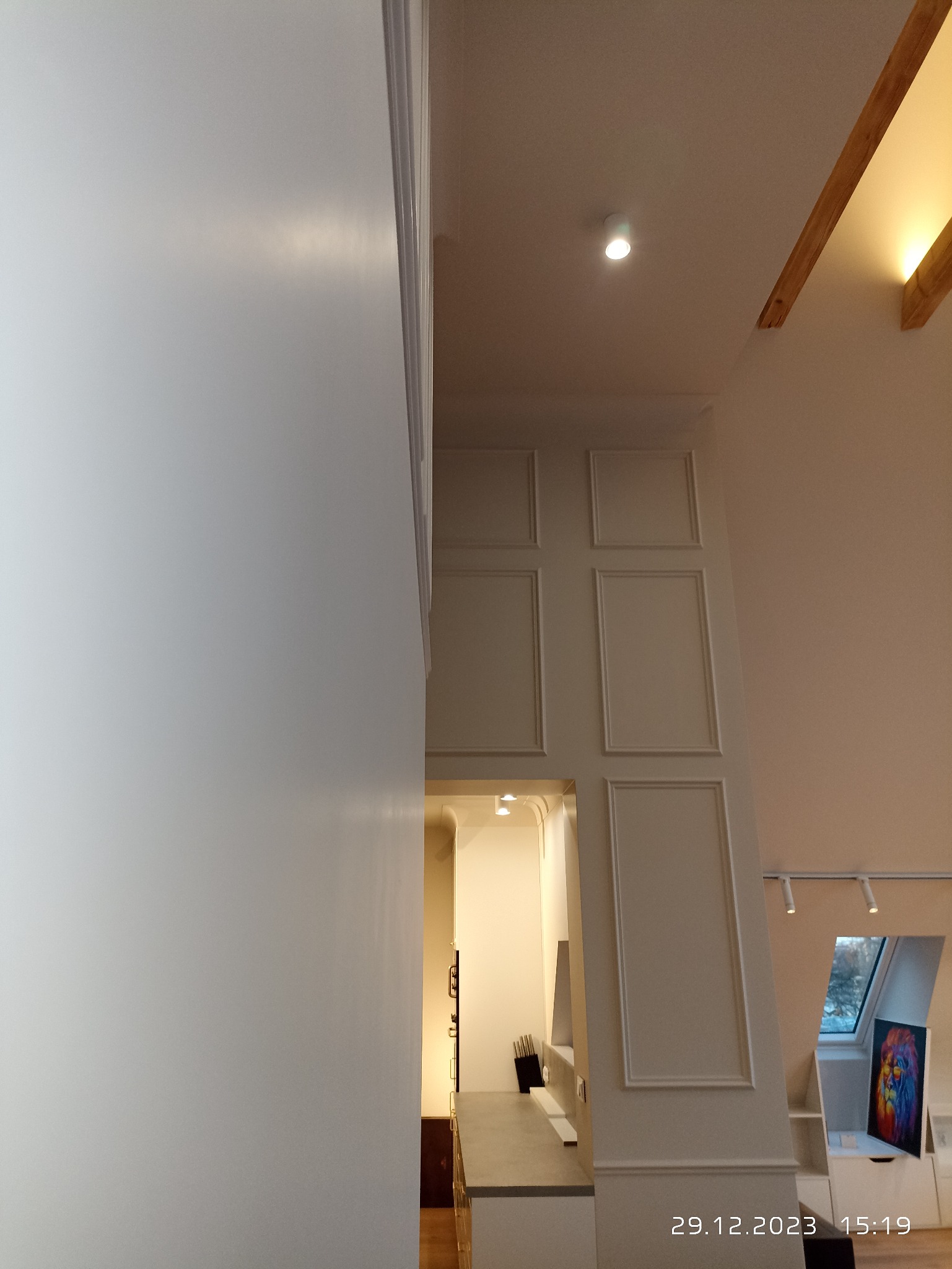 Wysokie pomieszczenie z białymi ścianami, sztukaterią i widokiem na kuchnię w głębi, oświetlone punktowo