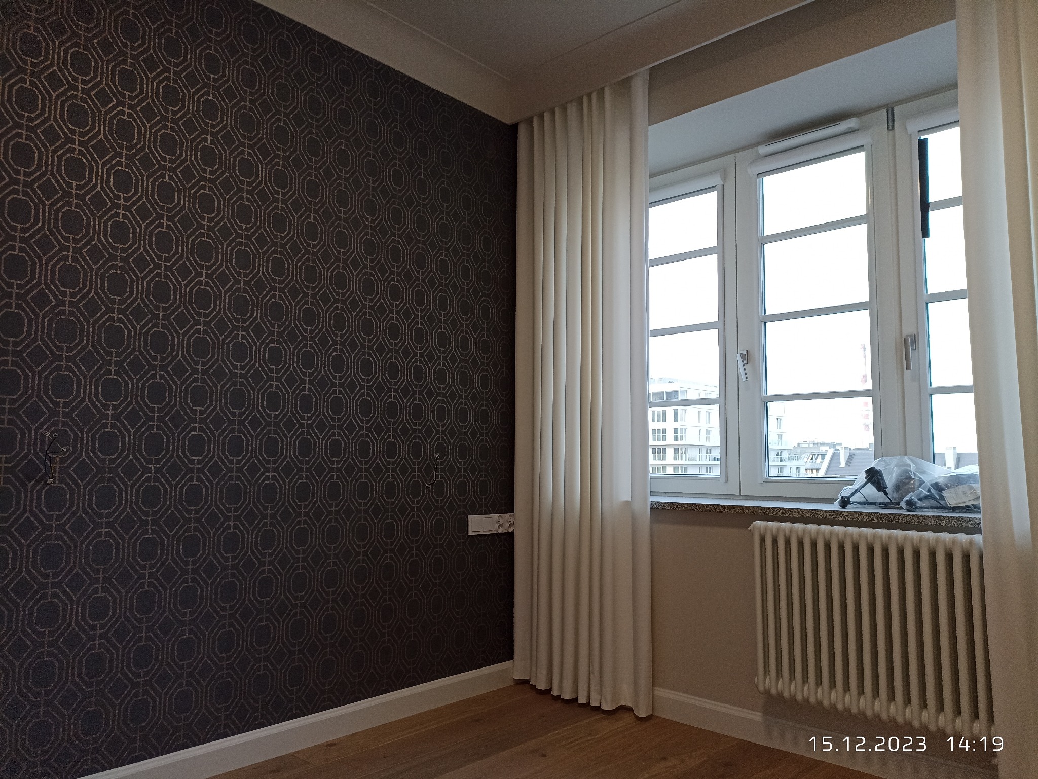 Pokój po odświeżeniu: ciemna ściana z geometrycznym wzorem, białe zasłony przy oknie z widokiem na miasto, grzejnik pod oknem, drewniana podłoga.