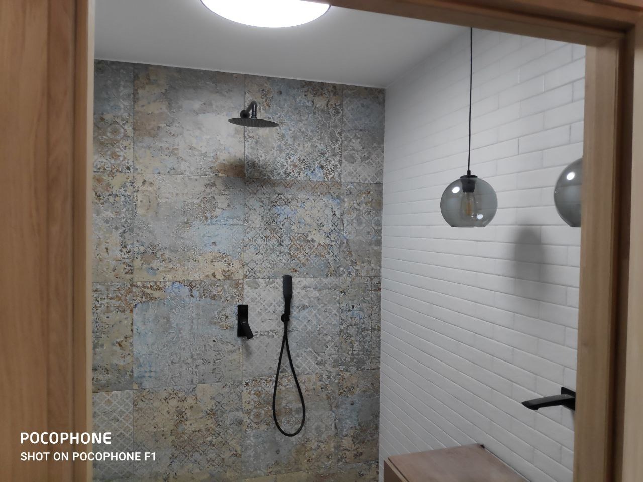 Nowoczesna kabina prysznicowa z deszczownicą i baterią w kolorze czarnym, płytki ścienne w stylu patchwork oraz białe, podłużne płytki, oświetlenie sufitowe w formie szklanych kul.