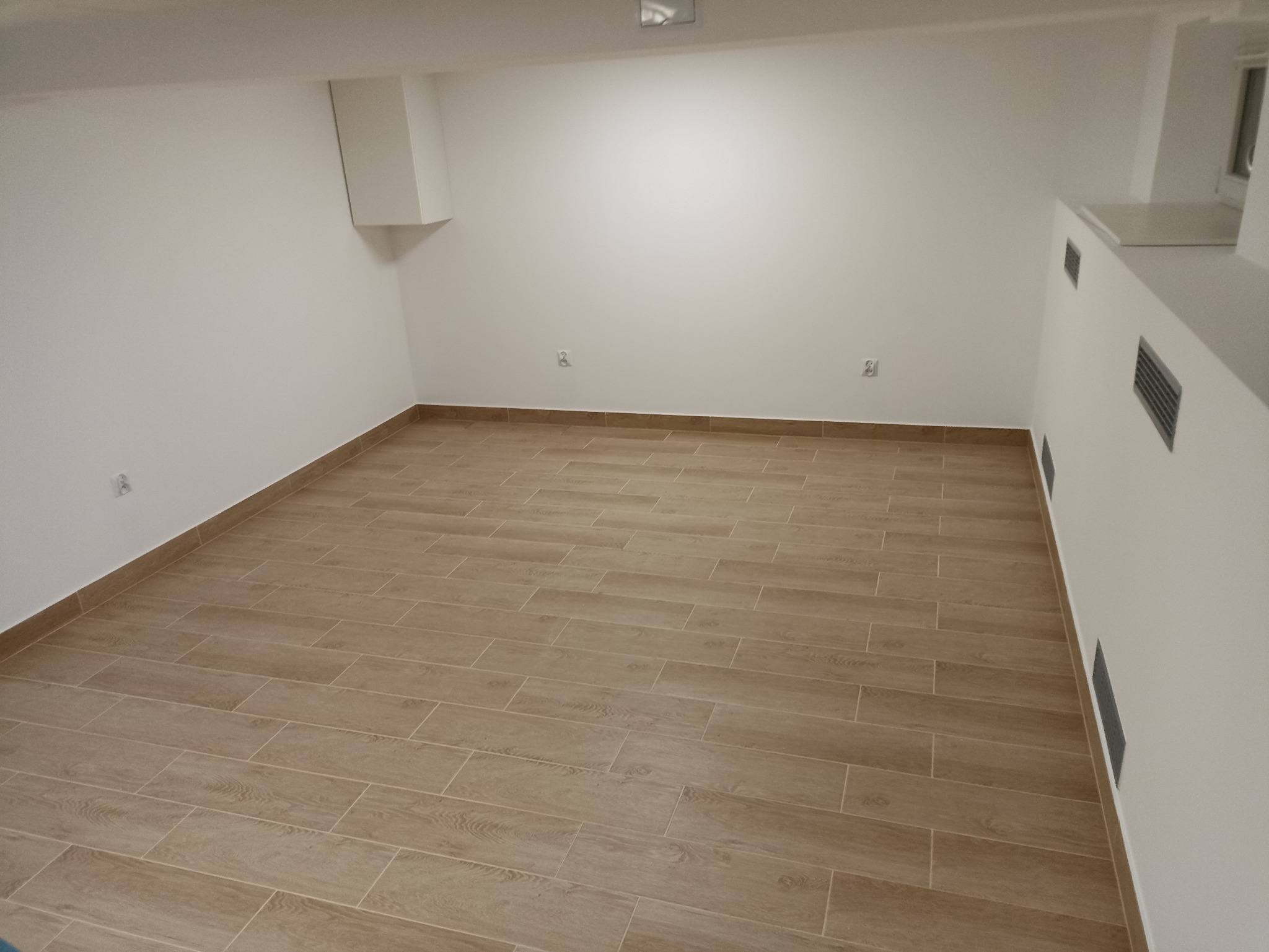 Puste pomieszczenie po ułożeniu podłogi z płytek imitujących drewno, widoczne białe ściany z gniazdkami i kratkami wentylacyjnymi.