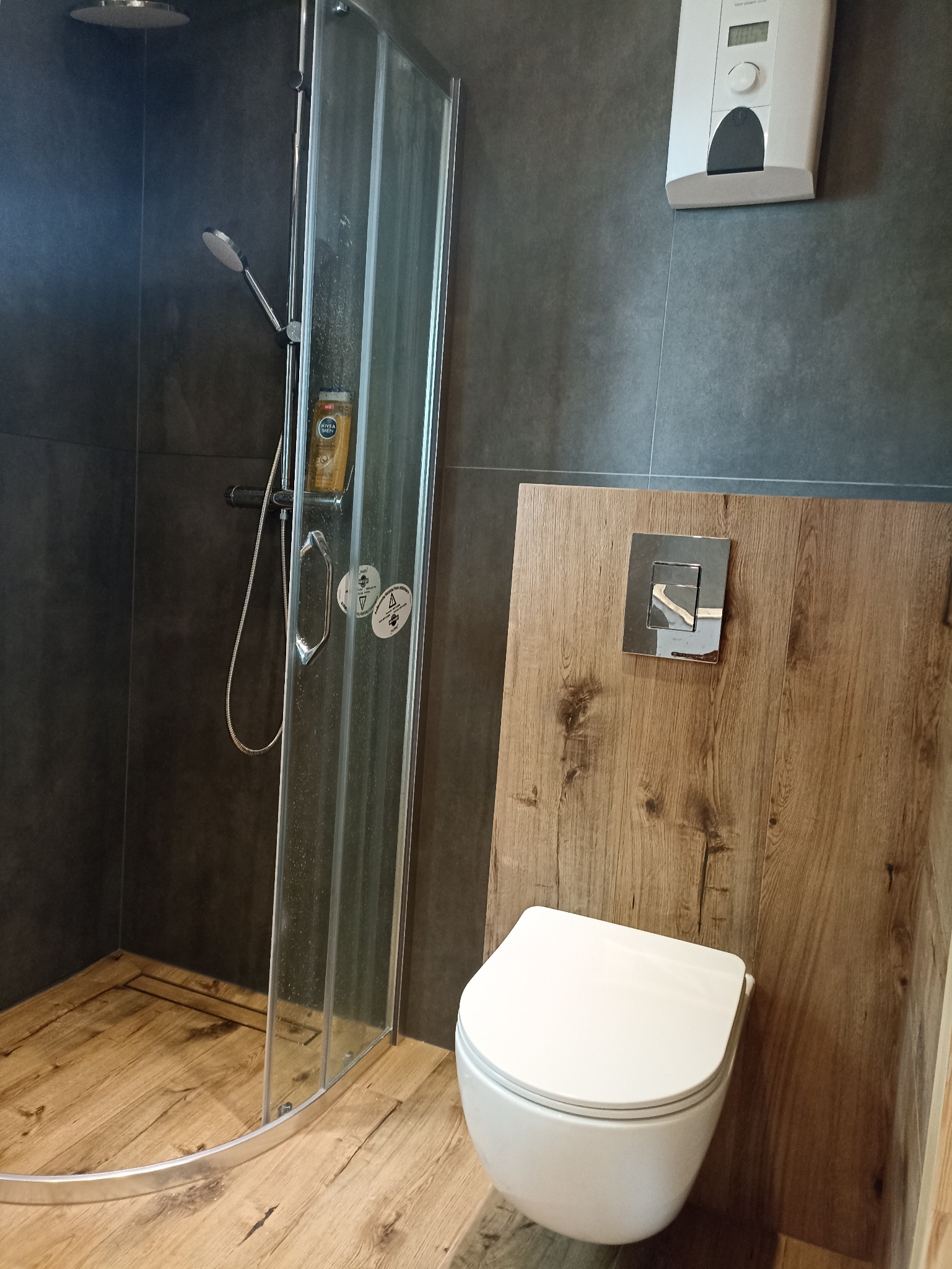 Nowoczesna łazienka z kabiną prysznicową z przezroczystego szkła, toaletą podwieszaną na drewnianej płycie i szarą ścianą z zamontowanym podgrzewaczem wody.