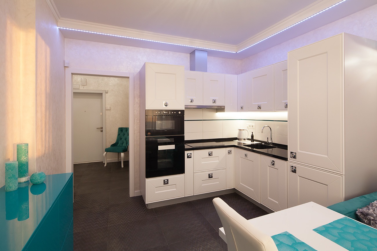 Biała, klasyczna zabudowa kuchenna z czarnym blatem, oświetleniem LED podszafkowym i górnym, widok na hol z turkusowym krzesłem i komodą, ciemna podłoga.