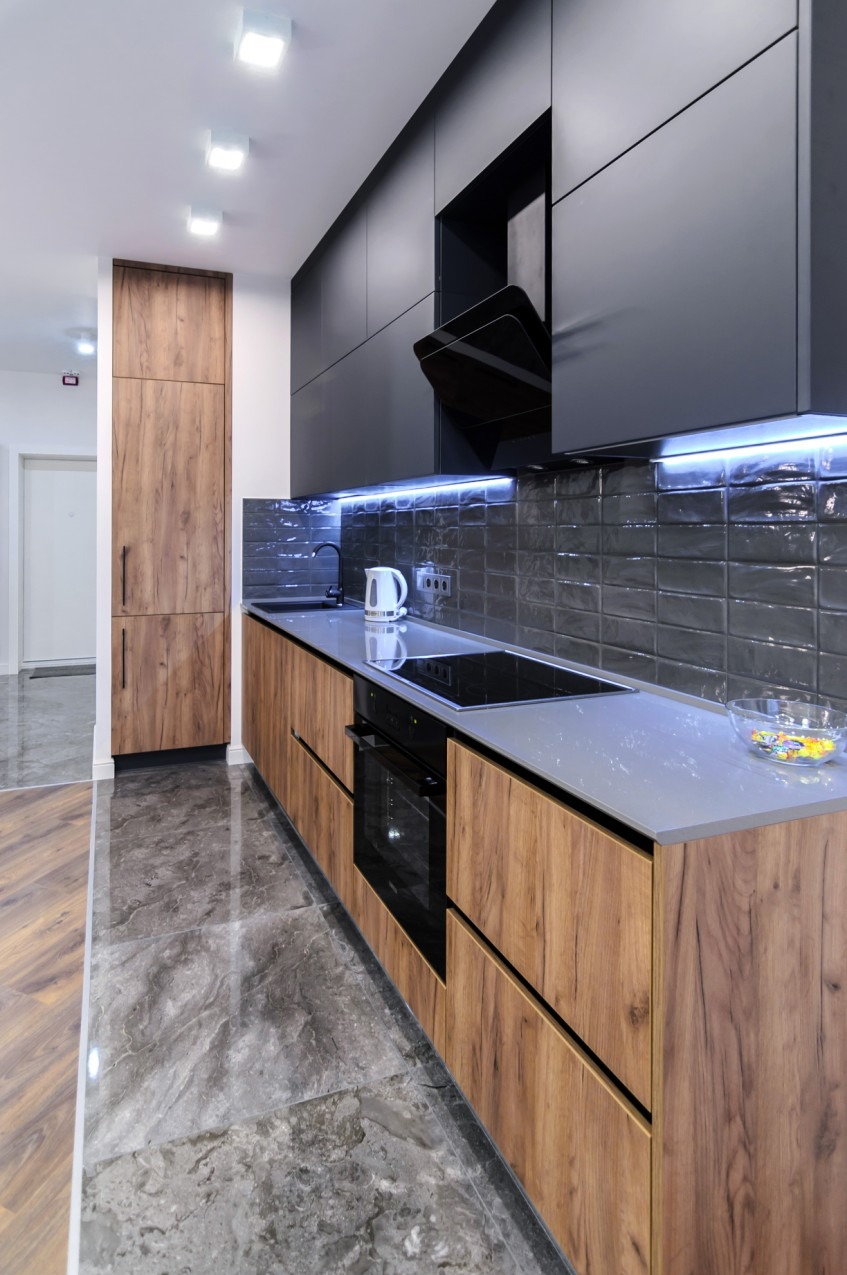 Nowoczesna kuchnia z drewnopodobnymi frontami szafek dolnych i czarnymi szafkami górnymi, szarym blatem i płytkami, oświetleniem LED pod szafkami wiszącymi i marmurową posadzką.