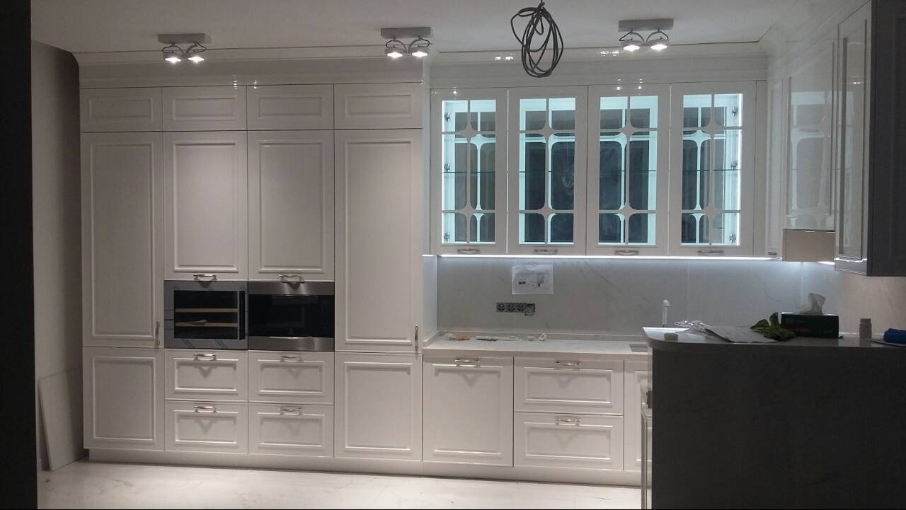 Klasyczna, biała zabudowa kuchenna z frezowanymi frontami, przeszklonymi szafkami górnymi i oświetleniem LED podszafkowym, w trakcie montażu; widoczne wystające kable i brakujące listwy przypodłogowe.