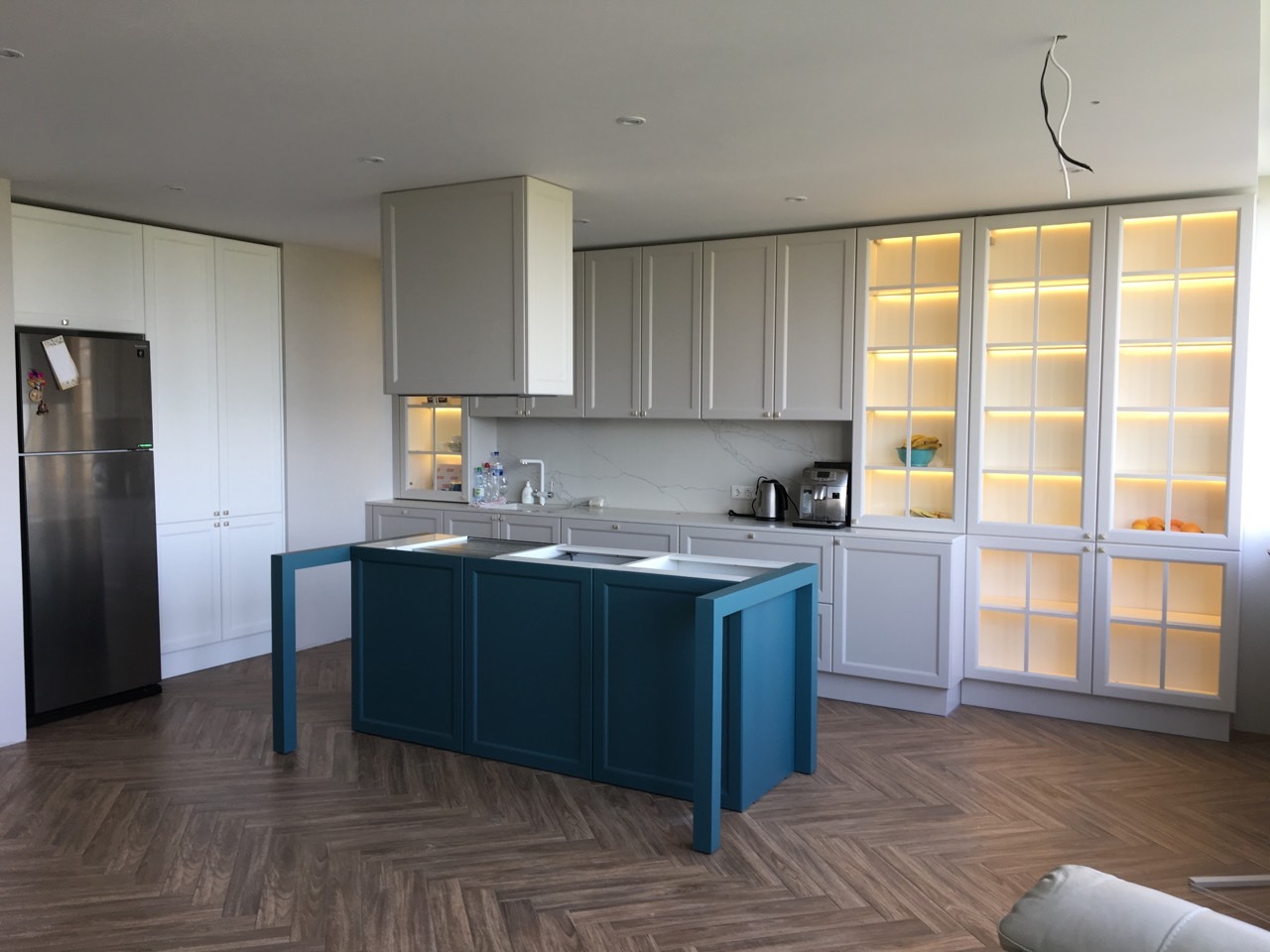 Nowoczesna kuchnia z wyspą w kolorze głębokiego błękitu, drewniana podłoga ułożona w jodełkę, białe szafki z podświetleniem, marmurowy backsplash, widoczne przewody elektryczne.