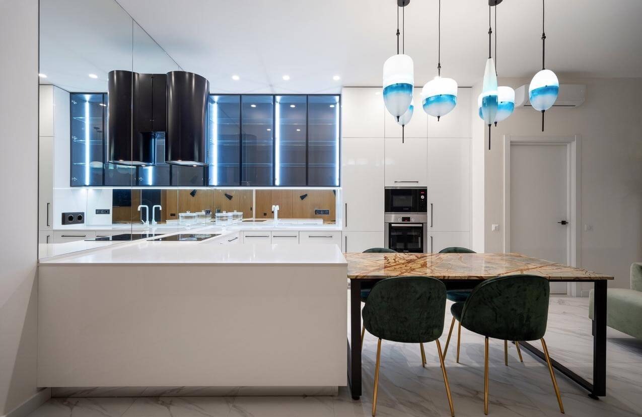 Nowoczesna kuchnia w bieli z wyspą, czarnym okapem o nietypowym kształcie i wiszącymi lampami z niebieskimi akcentami nad stołem z marmurowym blatem i zielonymi krzesłami.