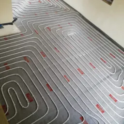 Białe rury ogrzewania podłogowego ułożone w pętle na szarej folii z czerwonym nadrukiem, widok z góry, częściowo widoczne ściany i okno.