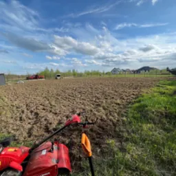Przygotowane pole pod trawnik w Koszalinie, widoczny glebogryzarka i szpadel, w tle zabudowa jednorodzinna i błękitne niebo z chmurami.