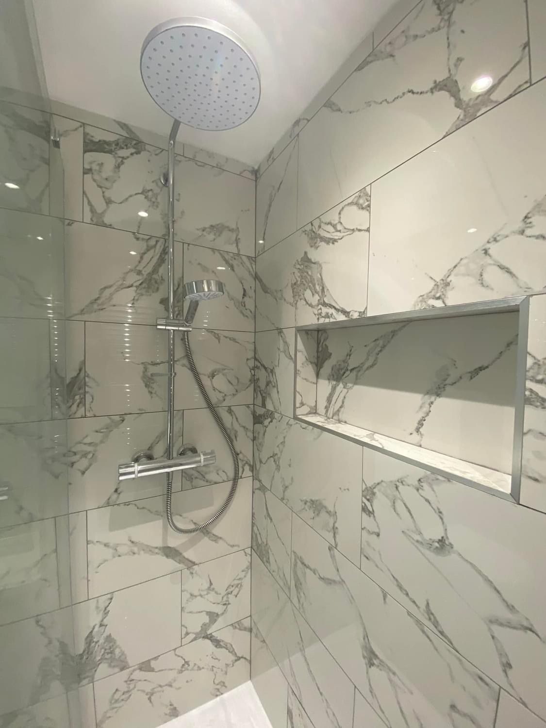 Kabina prysznicowa wykończona białymi płytkami imitującymi marmur z wnęką na kosmetyki, deszczownicą i baterią termostatyczną.