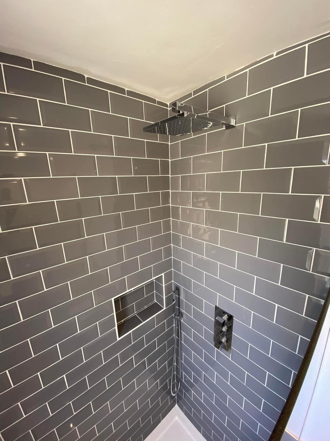 Nowoczesna kabina prysznicowa z szarymi, prostokątnymi płytkami ułożonymi horyzontalnie z białą fugą, deszczownicą i wnęką w ścianie.