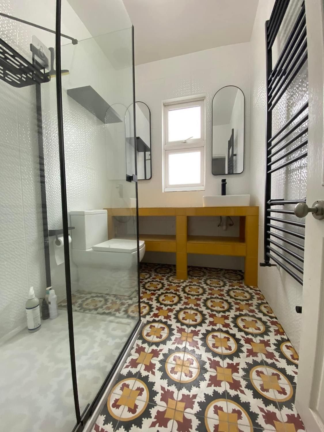 Nowoczesna łazienka z kabiną prysznicową typu walk-in, toaletą, żółtą szafką pod umywalką z czarną armaturą, dwoma owalnymi lustrami w czarnych ramach i grzejnikiem drabinkowym, na podłodze...