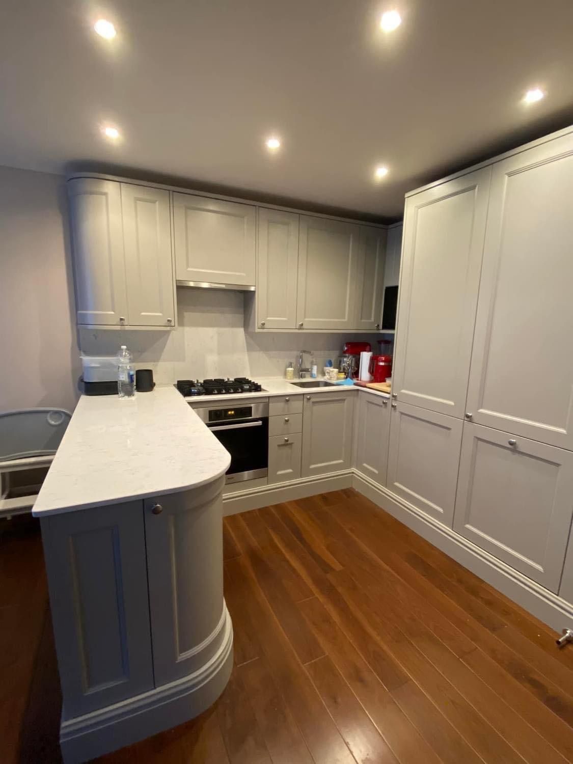 Jasnoszara kuchnia z drewnianą podłogą, wyspą z blatem imitującym marmur, zabudową do sufitu i oświetleniem punktowym.