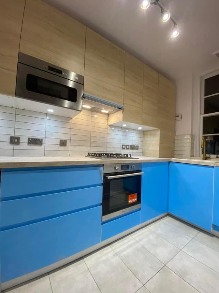 Nowoczesna kuchnia z jasnymi drewnianymi szafkami górnymi i niebieskimi dolnymi, płyta grzewcza nad piekarnikiem, białe płytki na ścianie, oświetlenie punktowe na suficie.