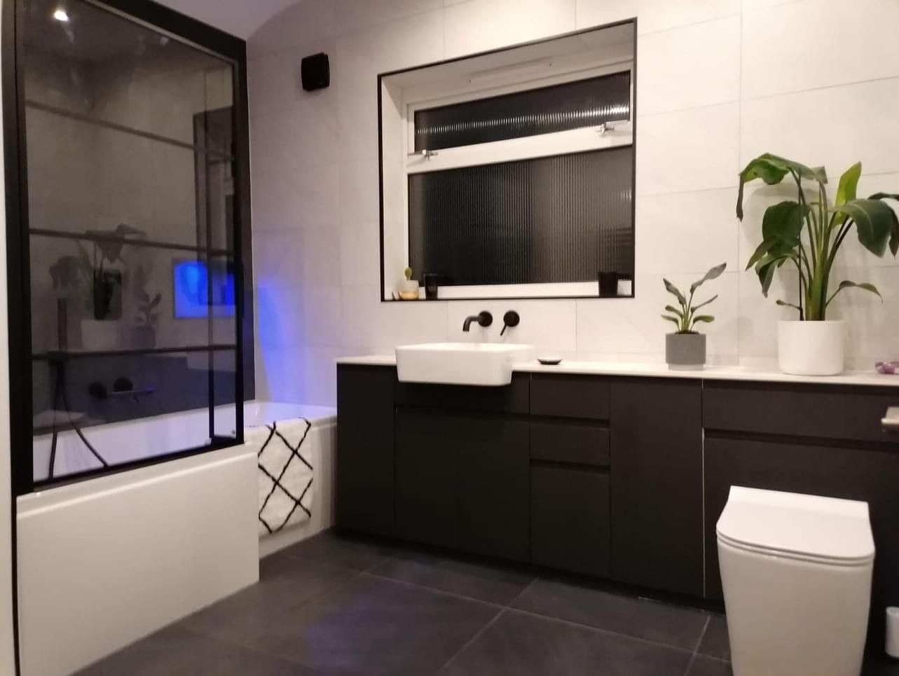 Nowoczesna łazienka z czarnymi akcentami: kabina prysznicowa z czarnymi ramami, umywalka nablatowa z czarną armaturą, duże lustro z czarną ramą, białe płytki na ścianach, ciemna podłoga, rośliny...