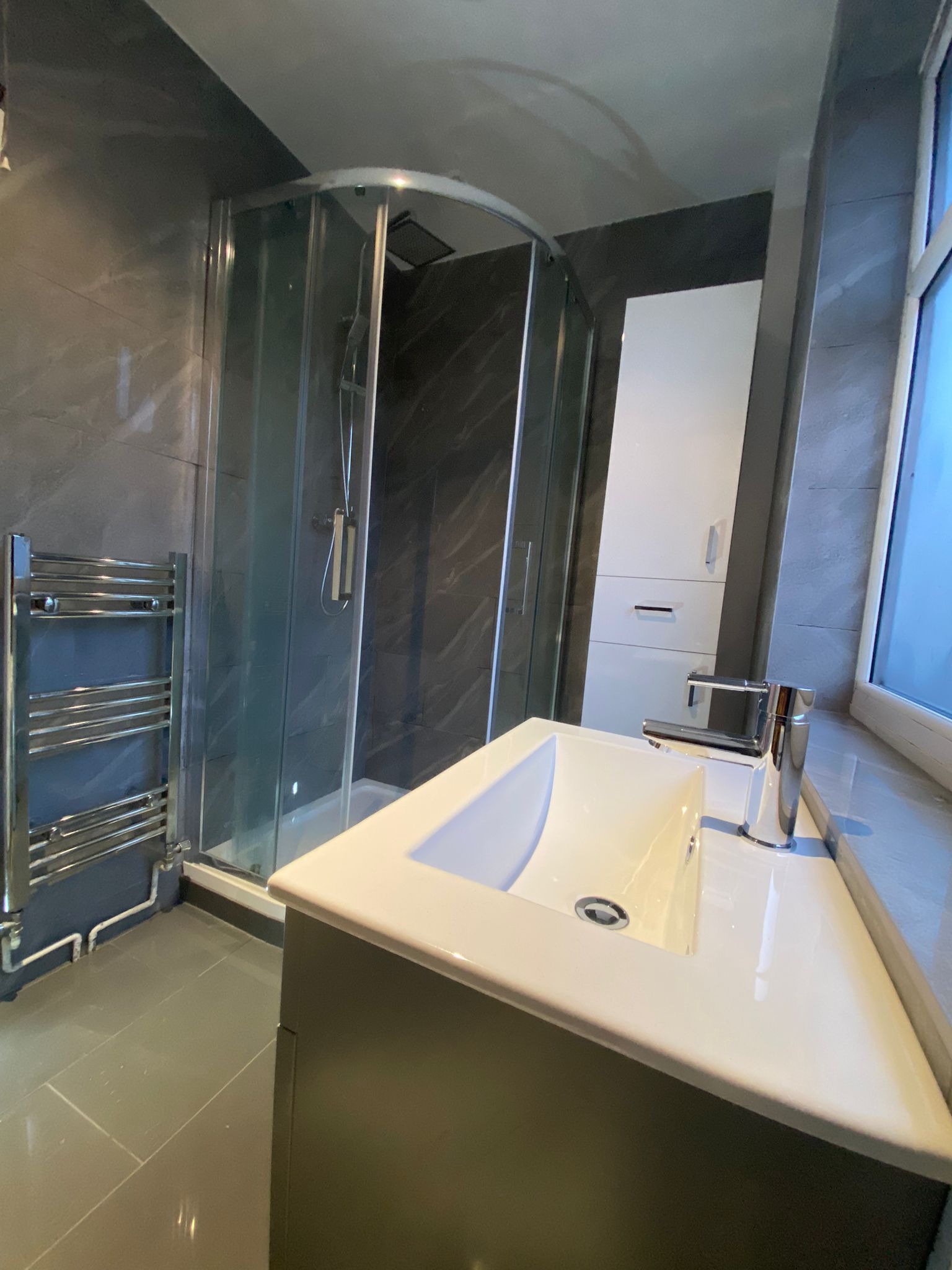 Nowoczesna łazienka z szarymi płytkami, kabiną prysznicową z zaokrąglonym szkłem, grzejnikiem drabinkowym i białą umywalką nablatową z chromowaną baterią.