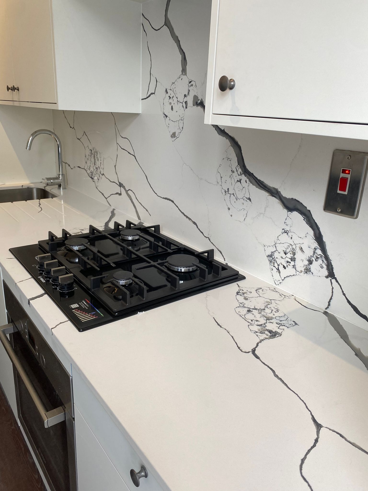 Biała kuchnia z blatem i ścianą z kwarcytu z wyraźnym czarnym żyłowaniem, czarna płyta gazowa wbudowana w blat, srebrny kran i włącznik światła.