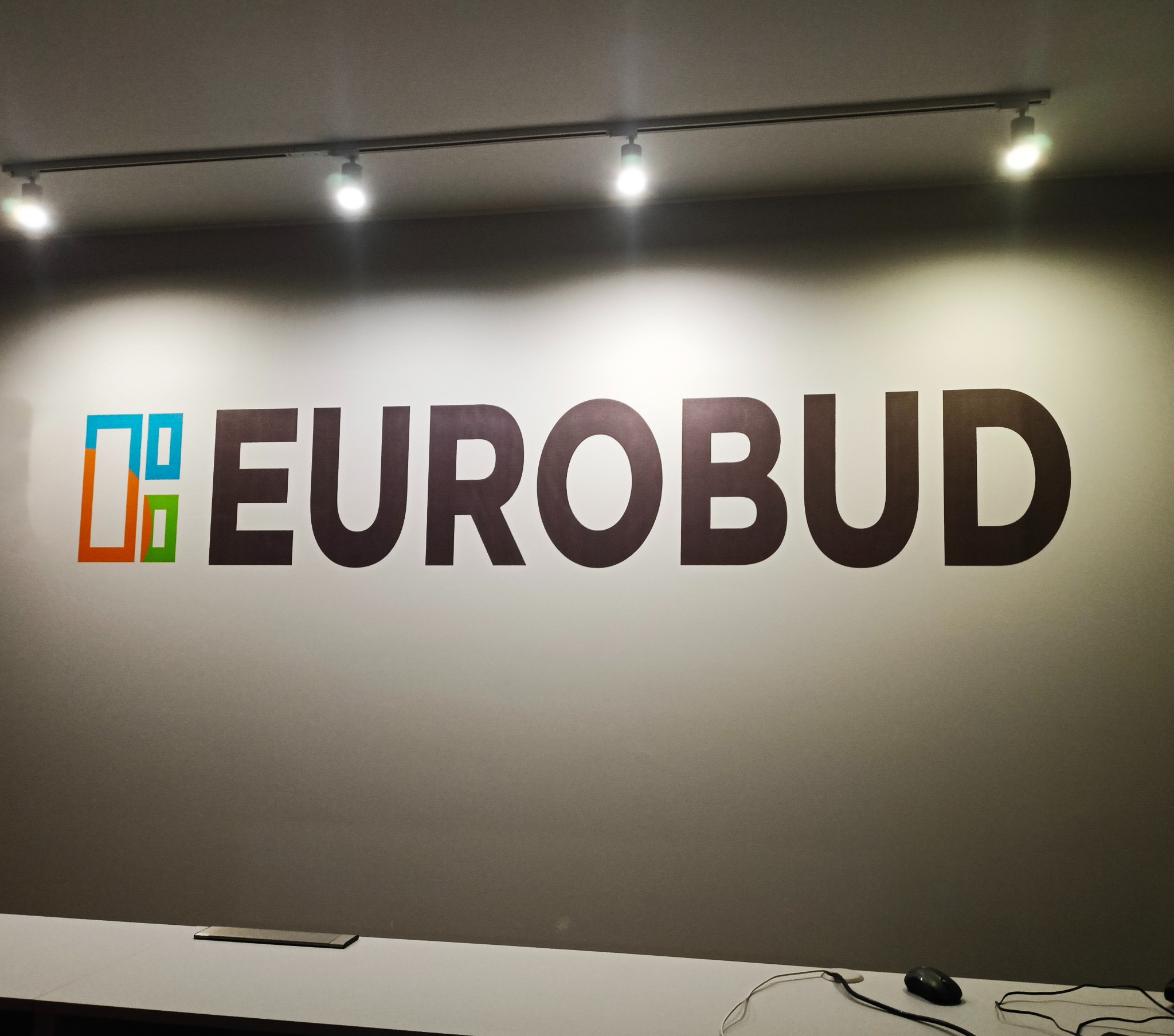 Ściana z logo firmy Eurobud, oświetlona reflektorami sufitowymi, z widocznym blatem biurka i myszką komputerową w dolnej części kadru.