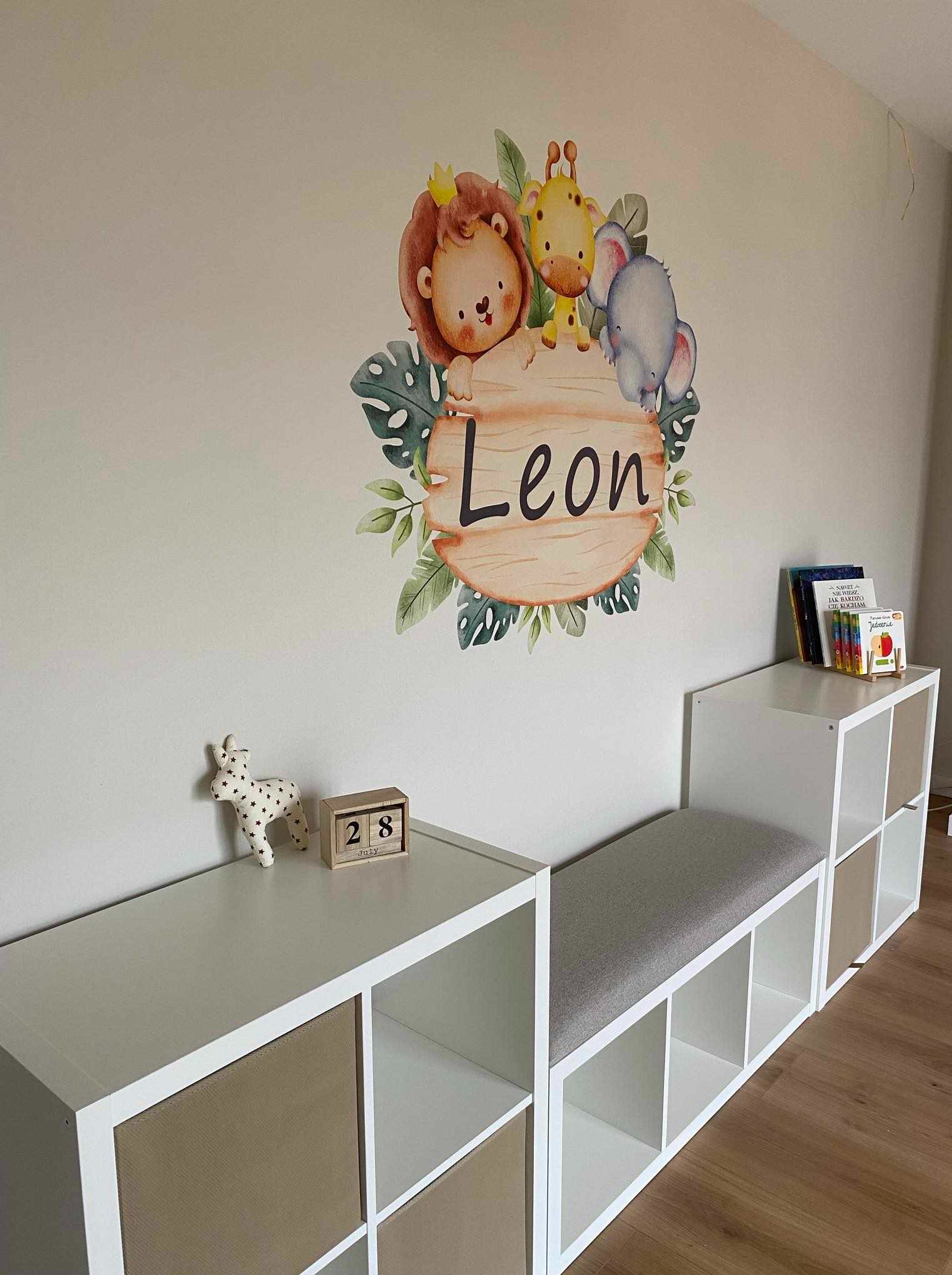 Pokój dziecięcy z naklejką ścienną przedstawiającą lwa w koronie, żyrafę i słonia z napisem 'Leon' na tle mebli modułowych.