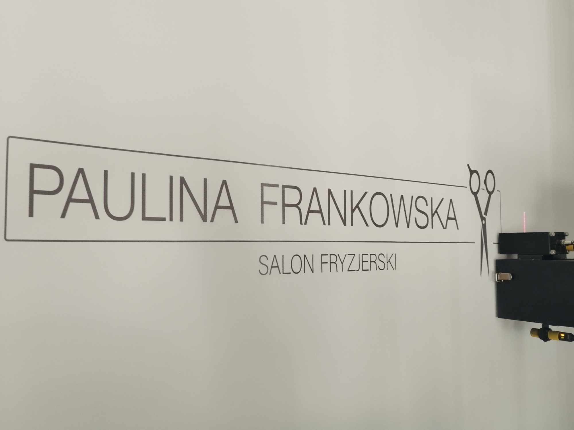 Czarny napis 'Paulina Frankowska Salon Fryzjerski' z symbolem nożyczek na ścianie w Gostyninie. Widoczny fragment urządzenia elektronicznego z kablami.