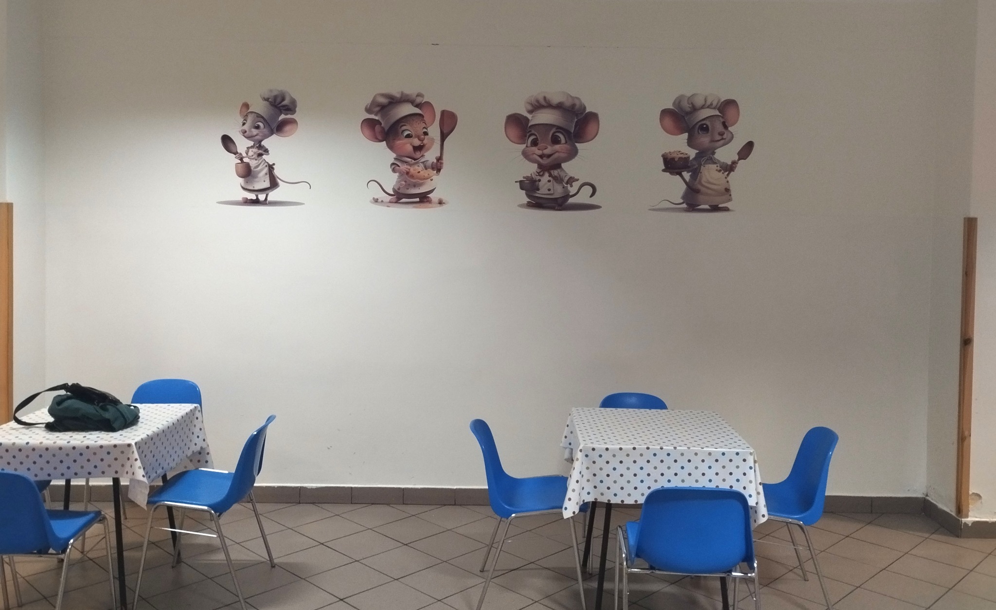 Ściana w jadalni z naklejkami czterech myszy-kucharzy w czapkach kucharskich, poniżej dwa stoliki nakryte obrusami w kropki i niebieskie krzesła.