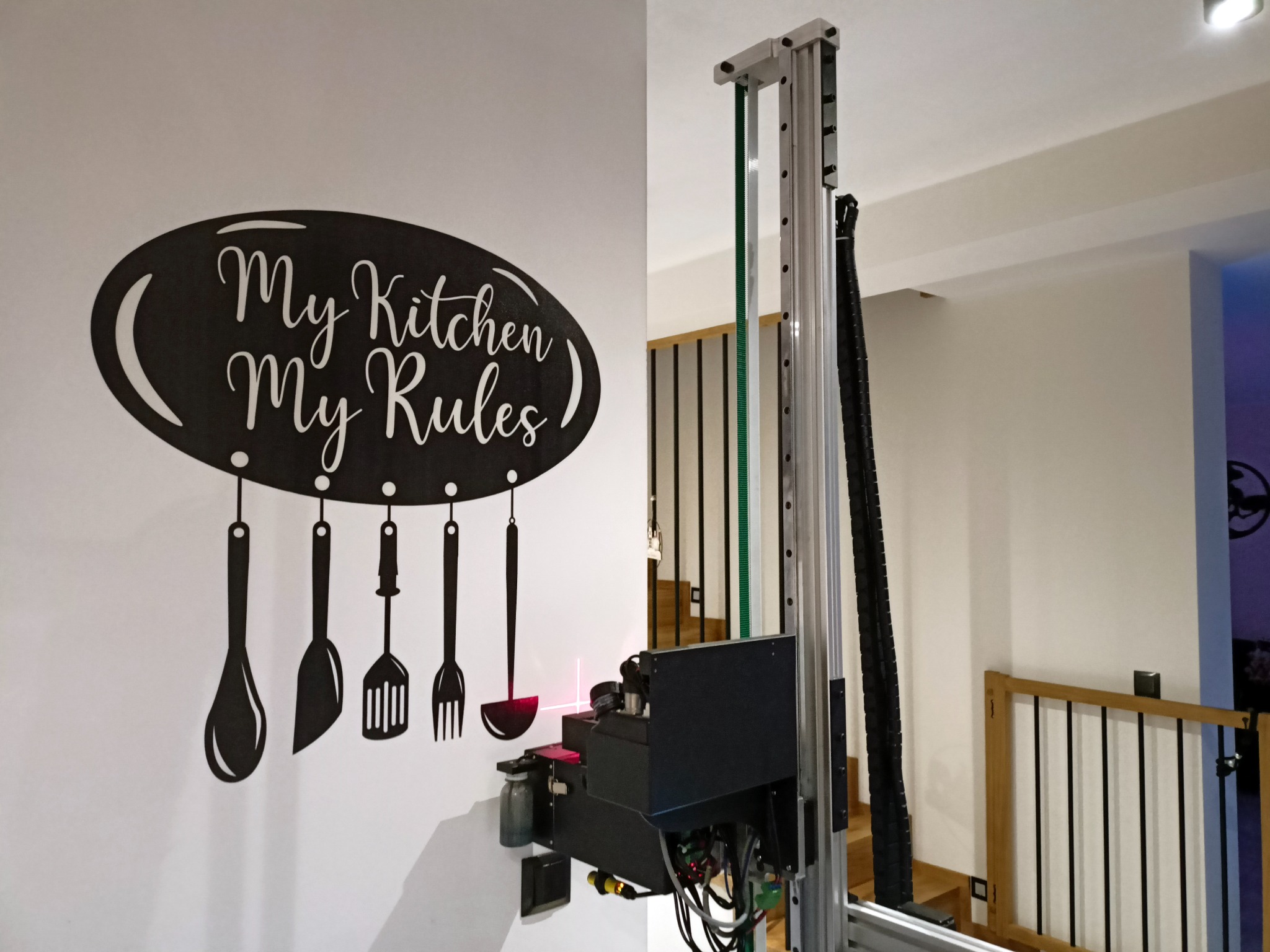 Drukarka pionowa nanosi czarny dekoracyjny napis 'My Kitchen My Rules' z grafiką przyborów kuchennych na białą ścianę w pomieszczeniu.