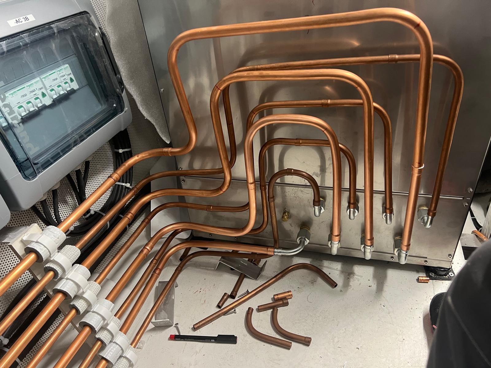Miedziane rury instalacji grzewczej, częściowo zamontowane, z widoczną skrzynką elektryczną i narzędziami na podłodze. Nietypowy układ rur.