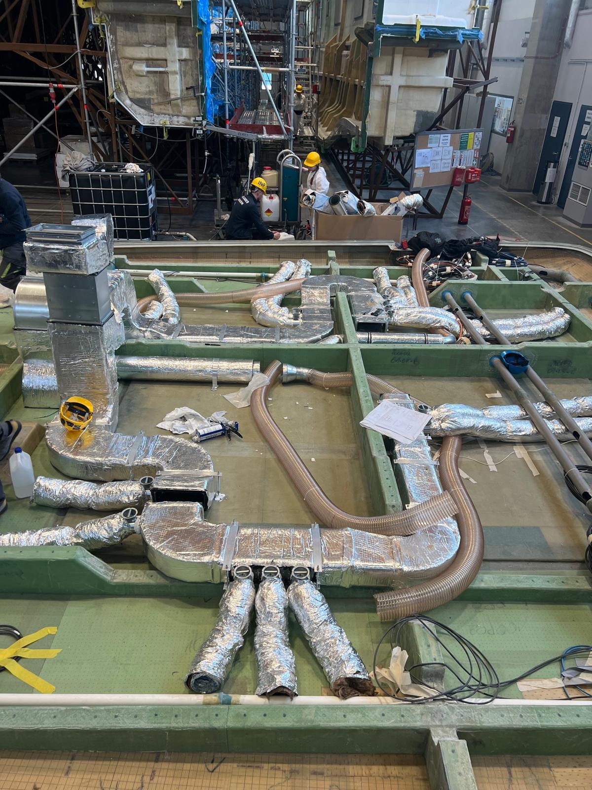 Instalacja wentylacyjna w trakcie montażu, rury owinięte folią aluminiową, widoczni pracownicy w kaskach, narzędzia pracy, zielona konstrukcja nośna.