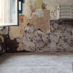 USŁUGI OGÓLNOBUDOWLANE PAWEŁ TELEGA - Ściana w klatce schodowej w trakcie remontu: widoczne warstwy starej farby, tynku i cegły. Okno z folią ochronną i skrzynki pocztowe. Etap przygotowania pod malowanie.