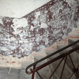 USŁUGI OGÓLNOBUDOWLANE PAWEŁ TELEGA - Przygotowanie ściany do malowania na klatce schodowej w Wałbrzychu. Widoczne ślady po szpachlowaniu i starej farbie, fragment schodów z metalową barierką.