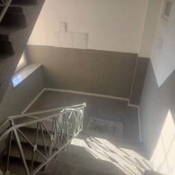 USŁUGI OGÓLNOBUDOWLANE PAWEŁ TELEGA - Klatka schodowa z szarymi ścianami o ziarnistej fakturze, białe szafki na ścianie i metalowa balustrada. Widok z góry na schody oświetlone naturalnym światłem.