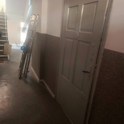 USŁUGI OGÓLNOBUDOWLANE PAWEŁ TELEGA - Klatka schodowa w trakcie malowania: drabina oparta o ścianę, pomalowane drzwi, osoba wchodząca po schodach. Widoczne ślady farby na podłodze.