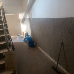 USŁUGI OGÓLNOBUDOWLANE PAWEŁ TELEGA - Klatka schodowa w trakcie malowania: widoczne wiadra z farbą, drabina, kartka z napisem 'Uwaga Malowane' i rysunkiem. Ściany pomalowane do połowy na szaro.