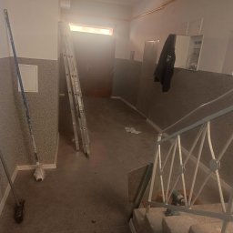 USŁUGI OGÓLNOBUDOWLANE PAWEŁ TELEGA - Klatka schodowa w trakcie odświeżania: drabina oparta o ścianę, wałek malarski, widoczne ślady prac malarskich. W tle drzwi i schody z metalową barierką.