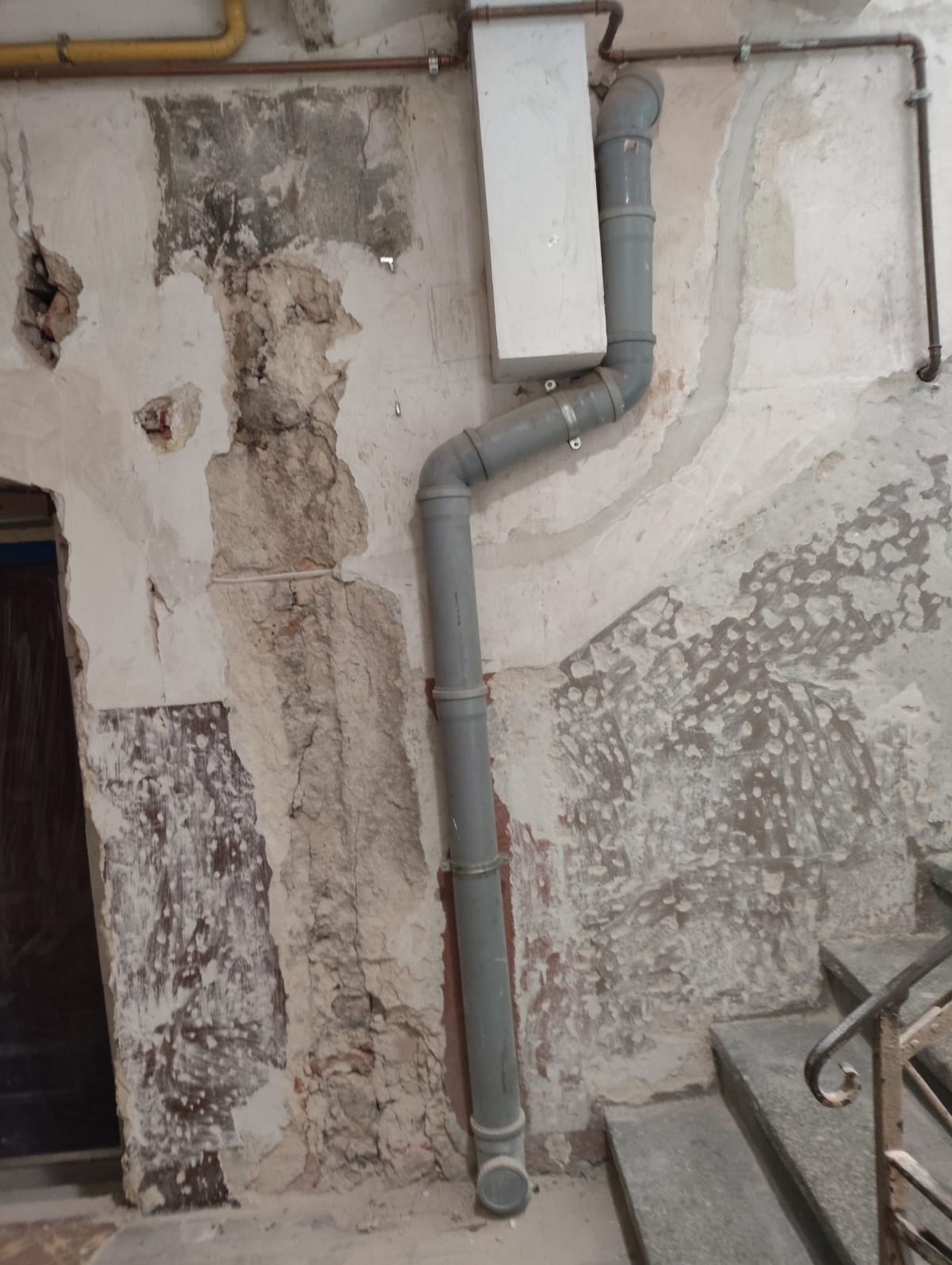 Fragment ściany w trakcie remontu z widoczną instalacją kanalizacyjną. Odsłonięta cegła i resztki starego tynku. Szare rury kanalizacyjne na tle surowej ściany.