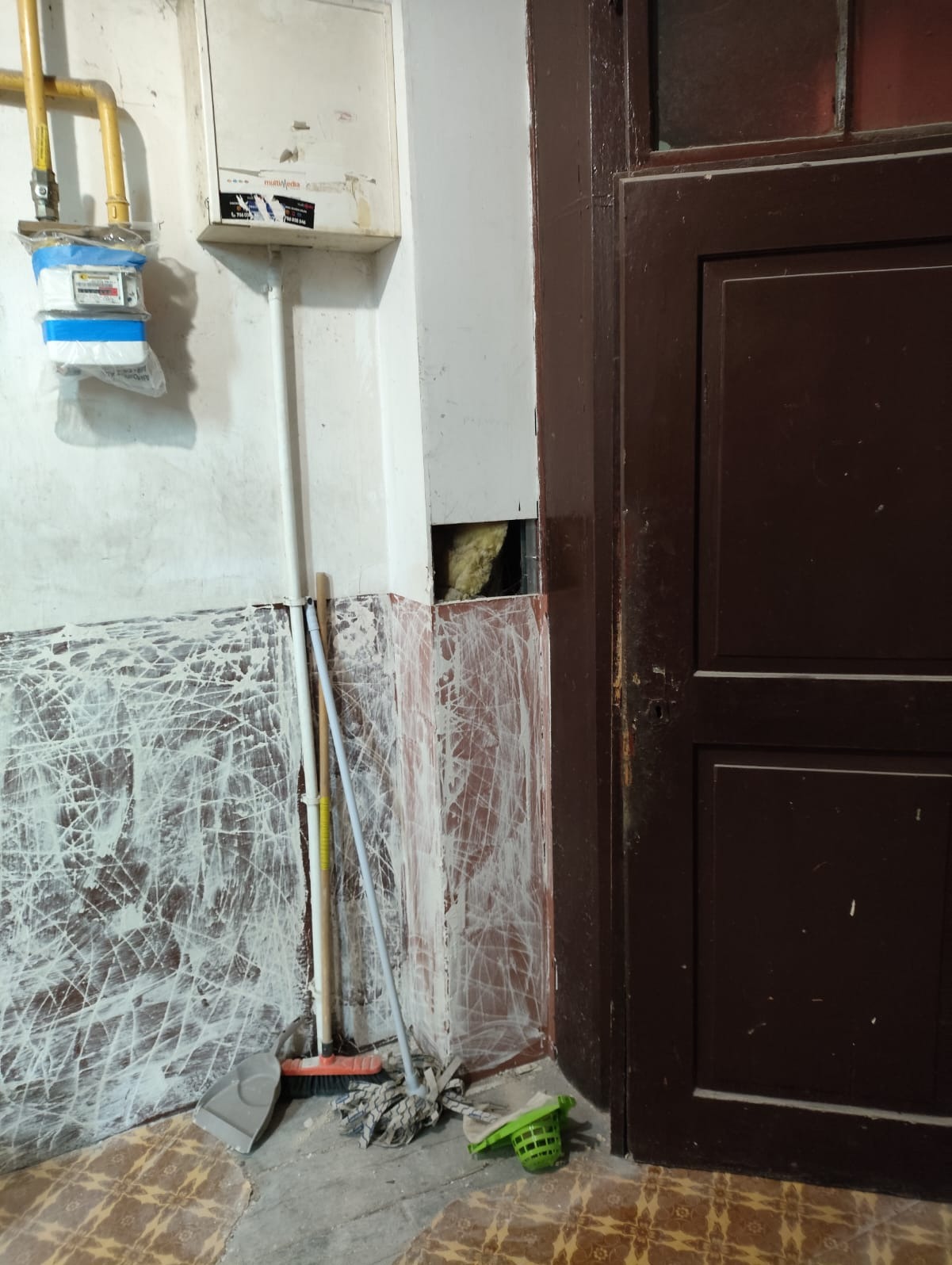 Fragment wnętrza w trakcie remontu: ściany z resztkami farby, odkryta izolacja w ścianie, narzędzia malarskie przy brązowych drzwiach. Widoczny gazomierz.