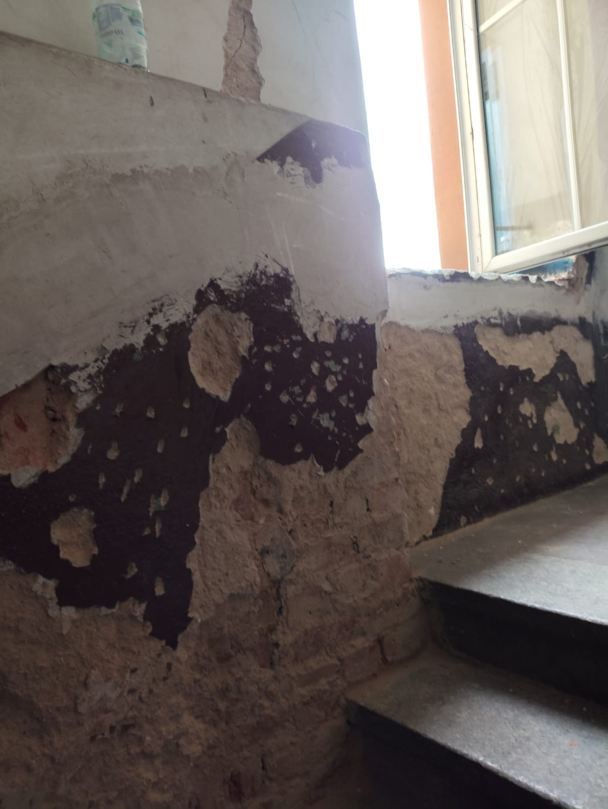 Odkryta cegła i resztki farby na ścianie przy schodach, widoczne ślady wcześniejszych prac remontowych, okno w tle.
