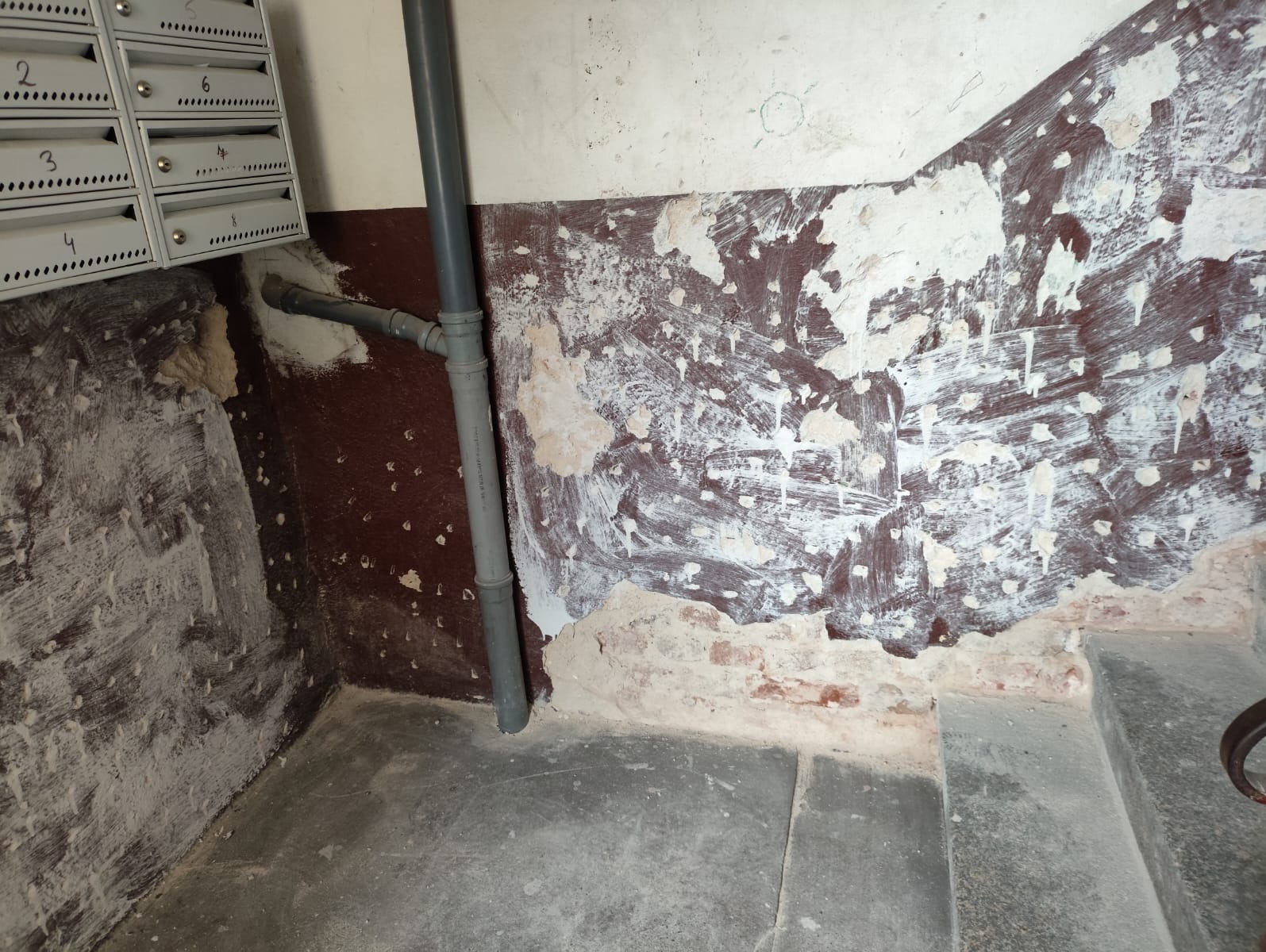Klatka schodowa w trakcie remontu: ściany z resztkami farby i gładzi, widoczne rury instalacyjne, skrzynki pocztowe. Etap przygotowania podłoża pod wykończenie.