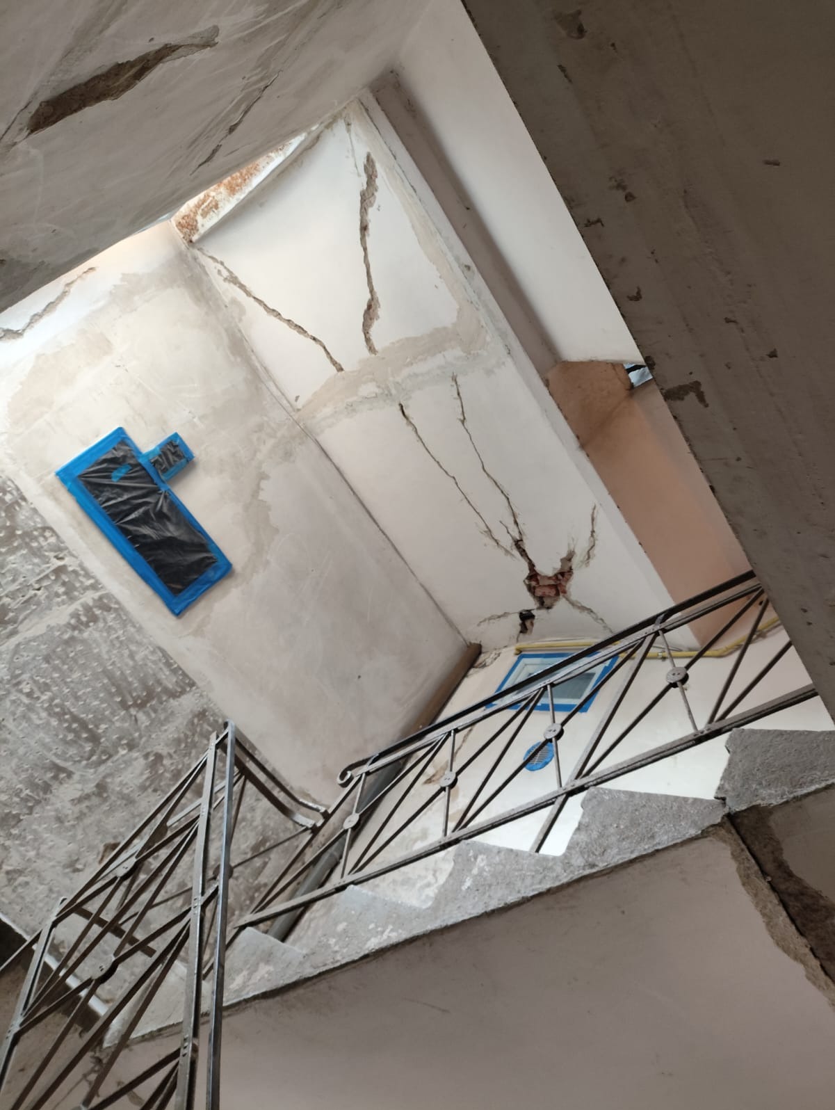 Wnętrze klatki schodowej w trakcie remontu: widoczne pęknięcia na ścianach i suficie, zabezpieczone okno folią, metalowa balustrada schodów. Ujęcie z dołu.