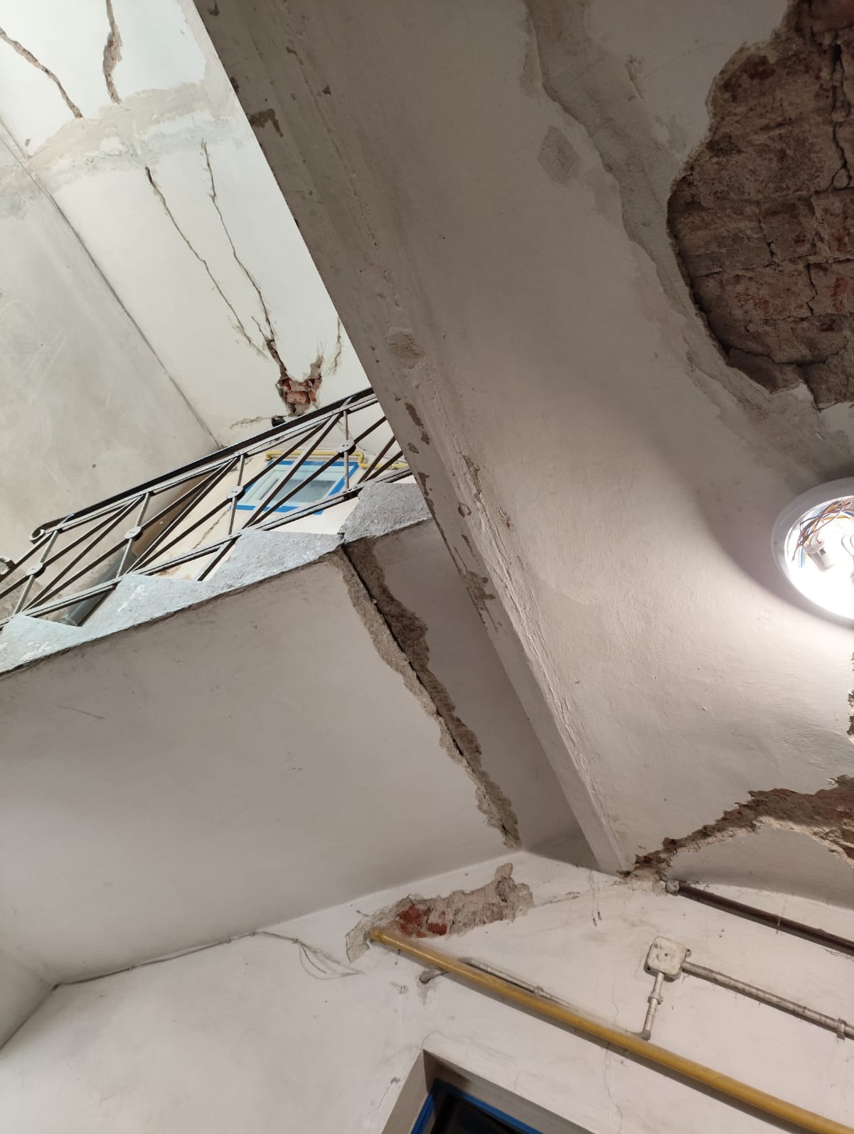 Uszkodzony sufit z pęknięciami i odsłoniętą cegłą, widok z dołu na klatce schodowej. Widoczne fragmenty balustrady i instalacji elektrycznej.